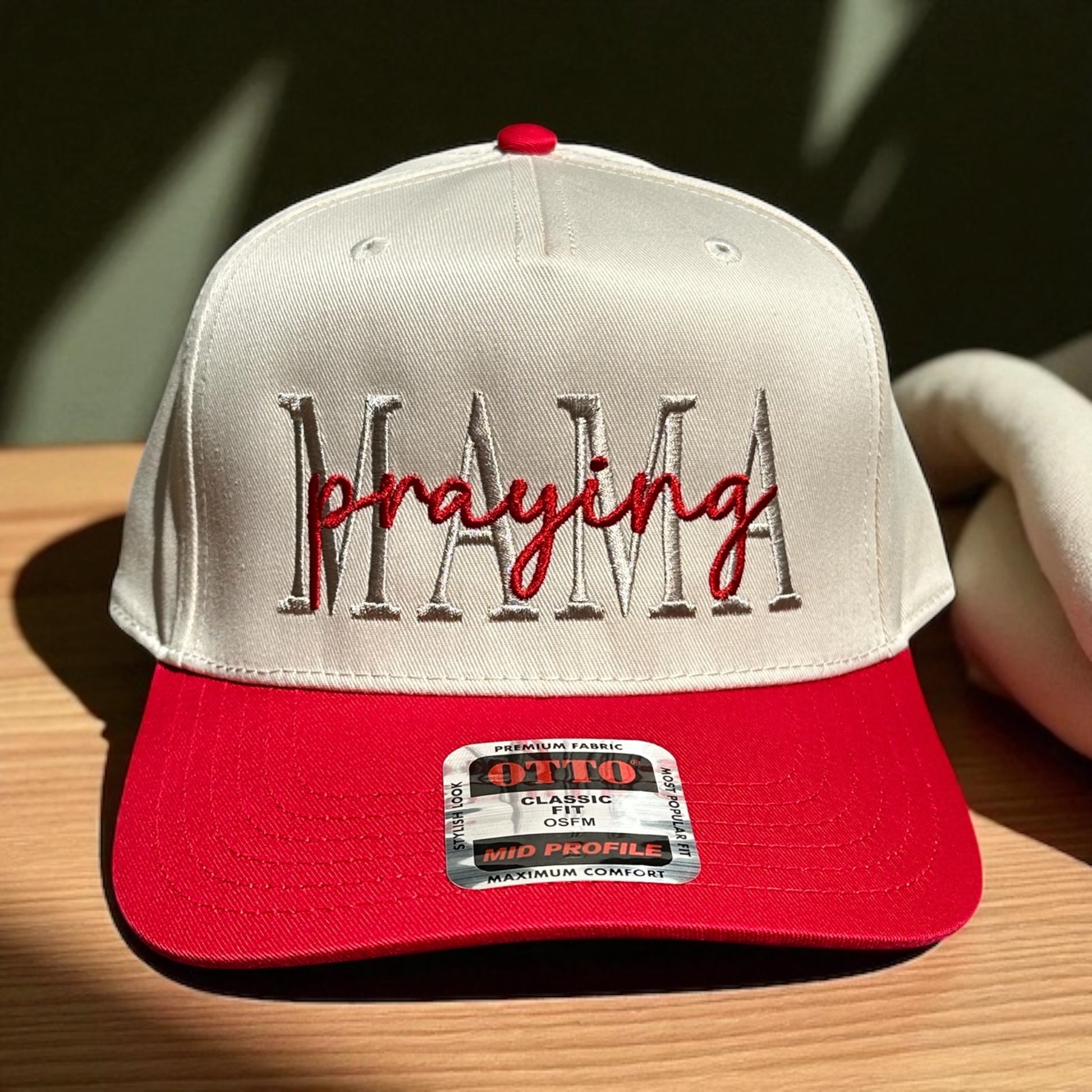 Two-tone cap with a light beige top and a red bill with embroidered text 'Praying Mama', and a visible brand logo label on a wooden surface and a dark green background