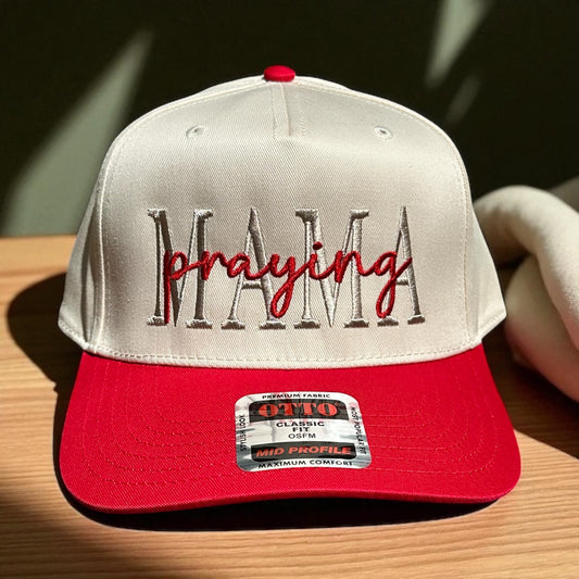 Two-tone cap with a light beige top and a red bill with embroidered text 'Praying Mama', and a visible brand logo label on a wooden surface and a dark green background