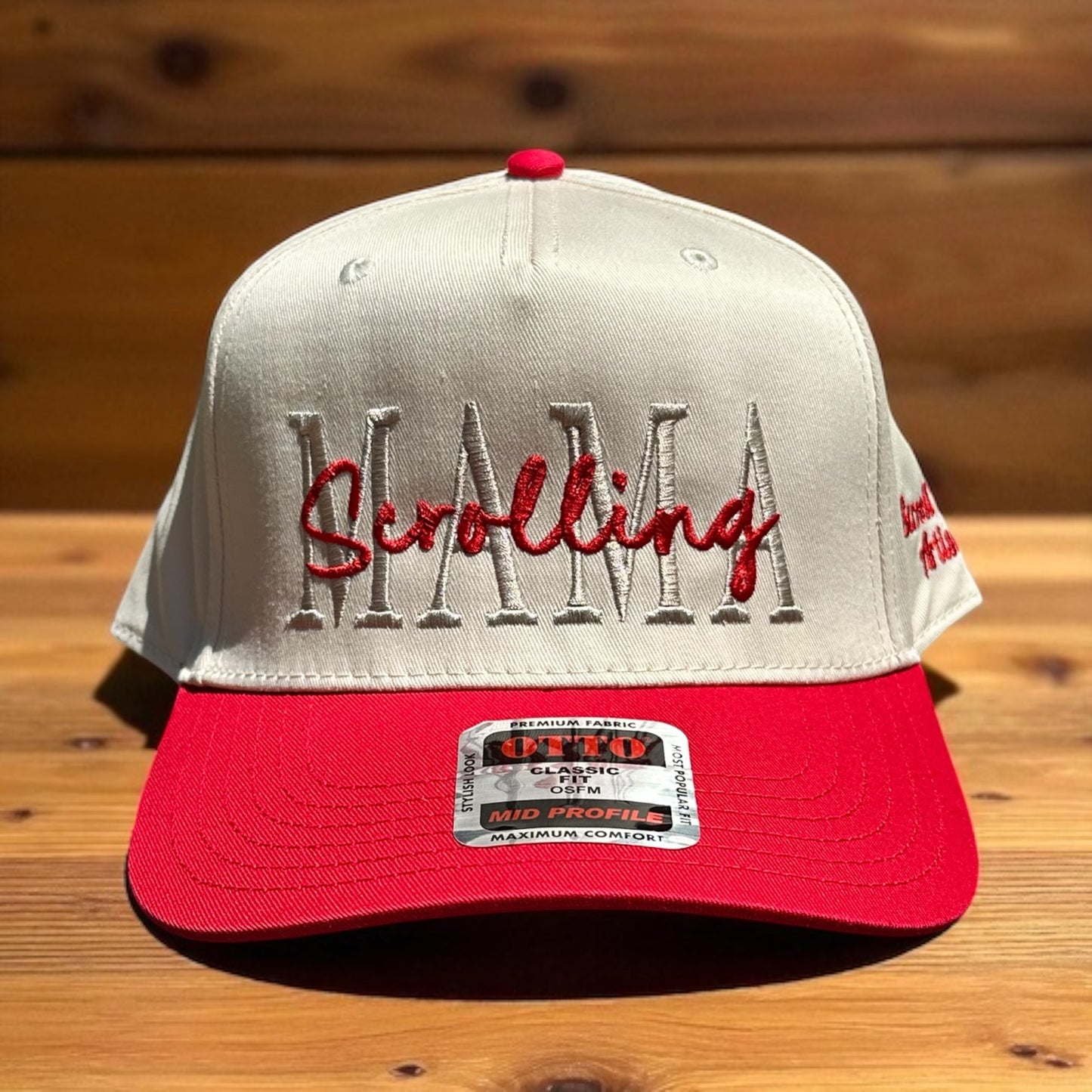 Two-tone cap with a light beige top and red bill with embroidered text 'Scrolling Mama', and a visible brand logo label, on a wooden surface