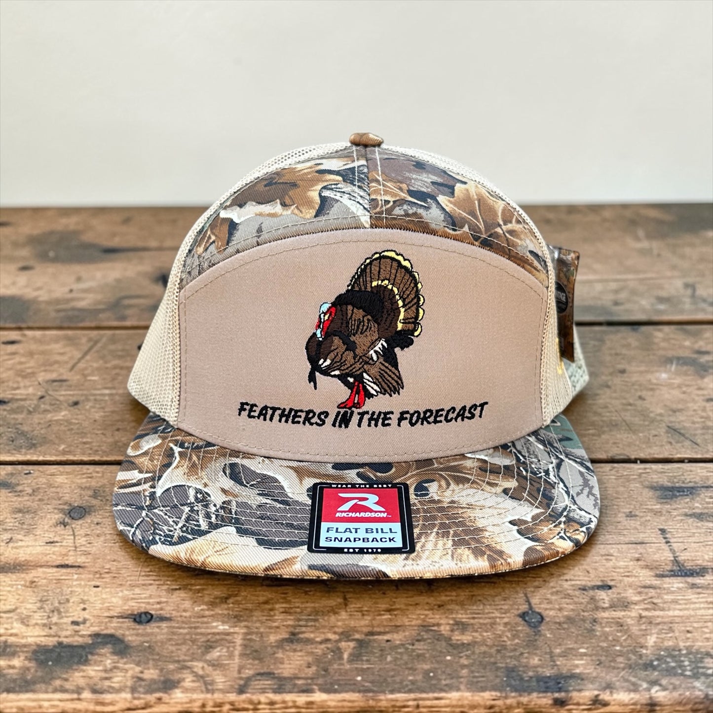 Camouflage and beige trucker cap with an embroidered turkey design and 'Feathers in the Forecast' text on a wooden surface.