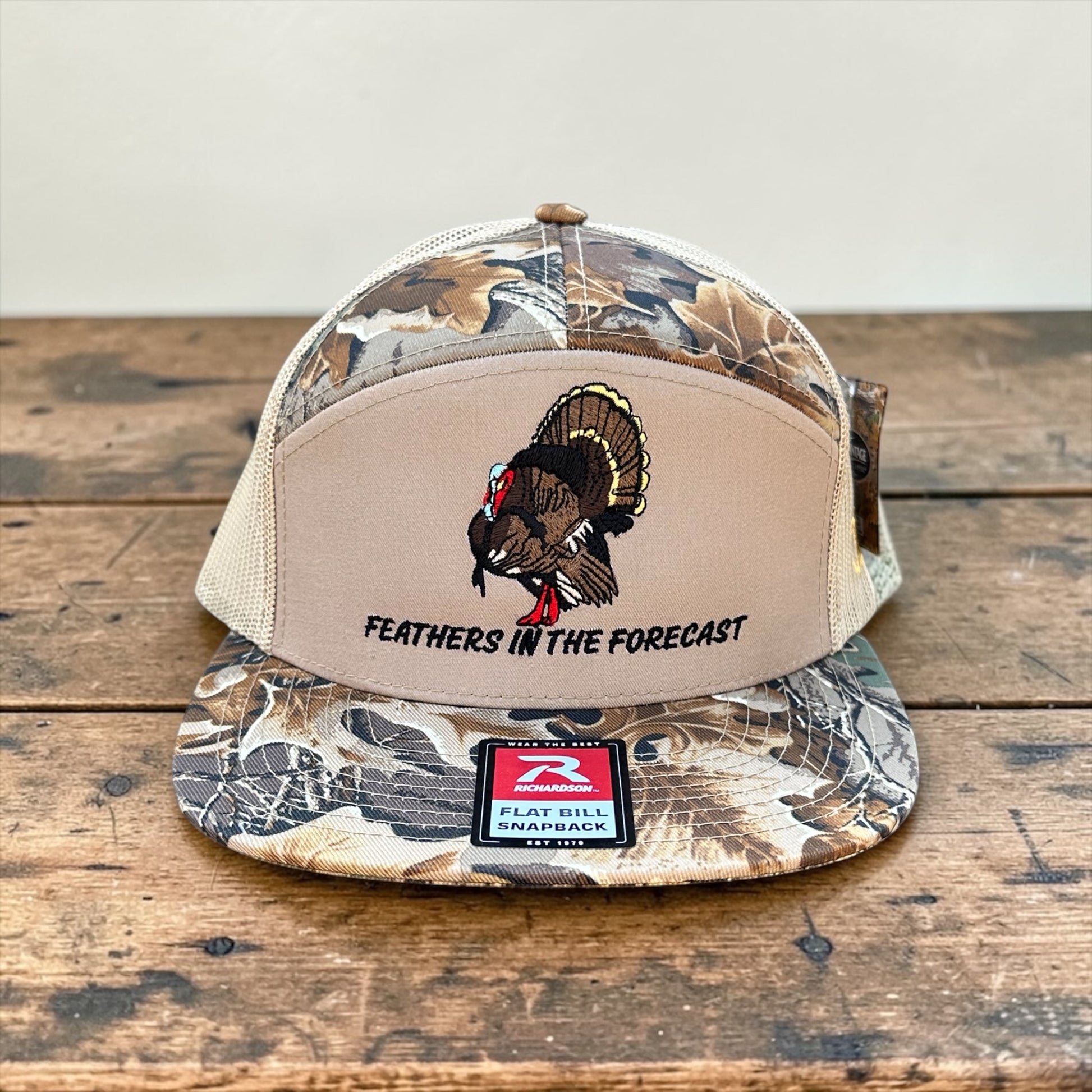 Camouflage and beige trucker cap with an embroidered turkey design and 'Feathers in the Forecast' text on a wooden surface.