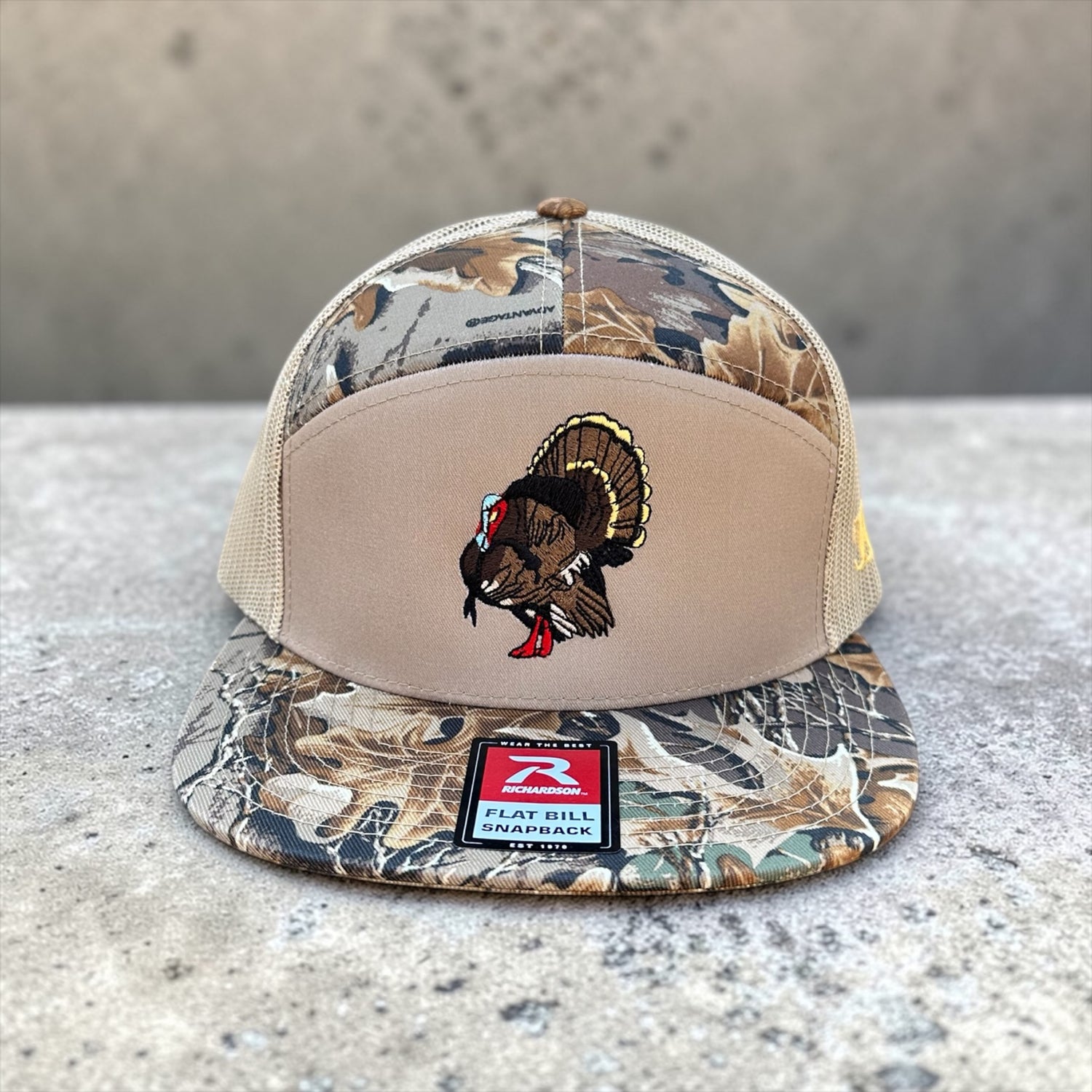Camouflage and beige trucker cap with an embroidered turkey design on a concrete surface
