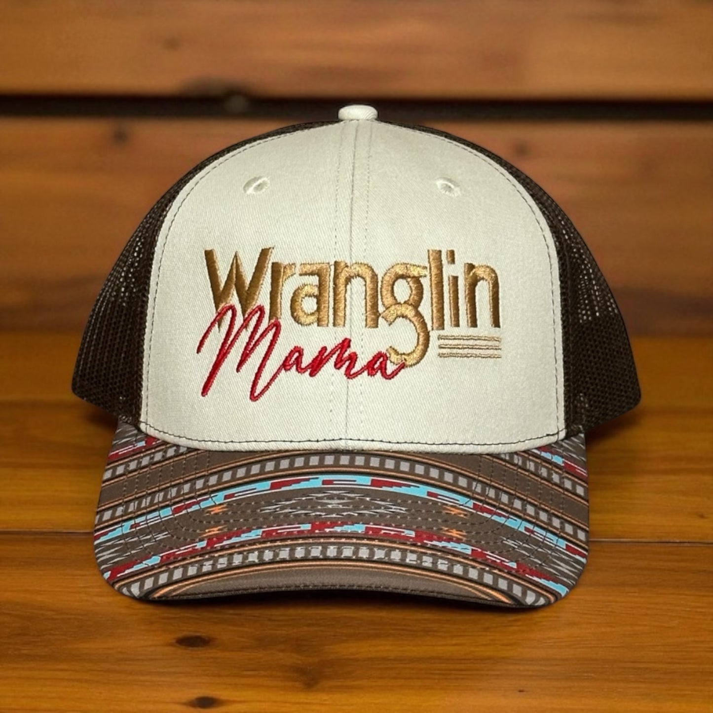 Tri-color trucker cap with a beige top, brown mesh back, and a multi-color southwestern pattern bill, with embroidered text 'Wranglin Mama', on a wooden surface