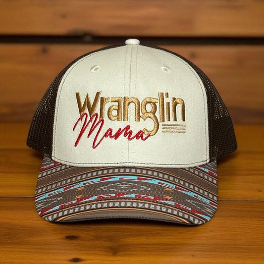 Tri-color trucker cap with a beige top, brown mesh back, and a multi-color southwestern pattern bill, with embroidered text 'Wranglin Mama', on a wooden surface