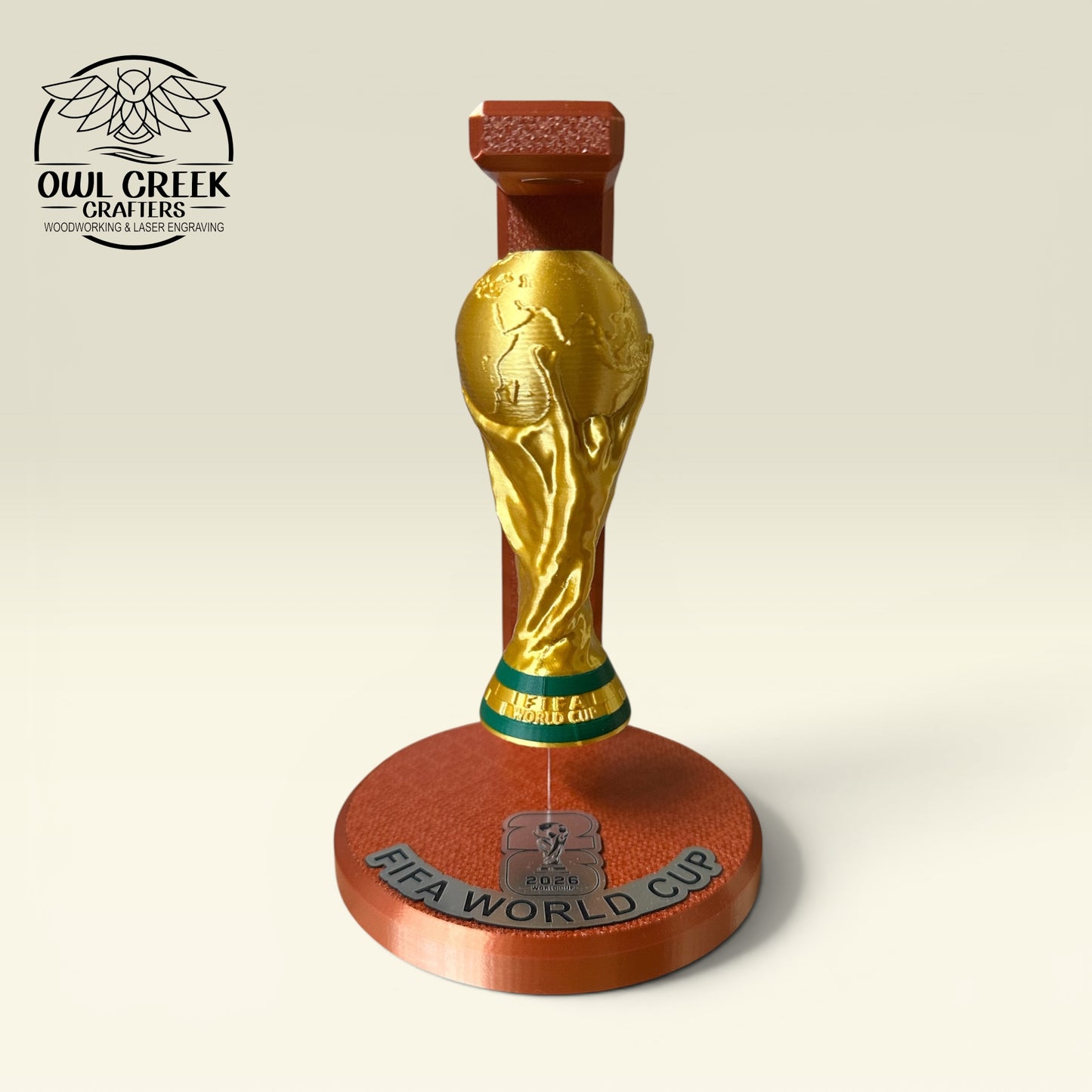 A 3d printed replica of the FIFA World Cup trophy on a 3d printed stand, featuring a gold-colored globe and a bronze copper base with 'FIFA WORLD CUP' text, shown on a light beige background with the Owl Creek Crafters logo in the top-left corner.