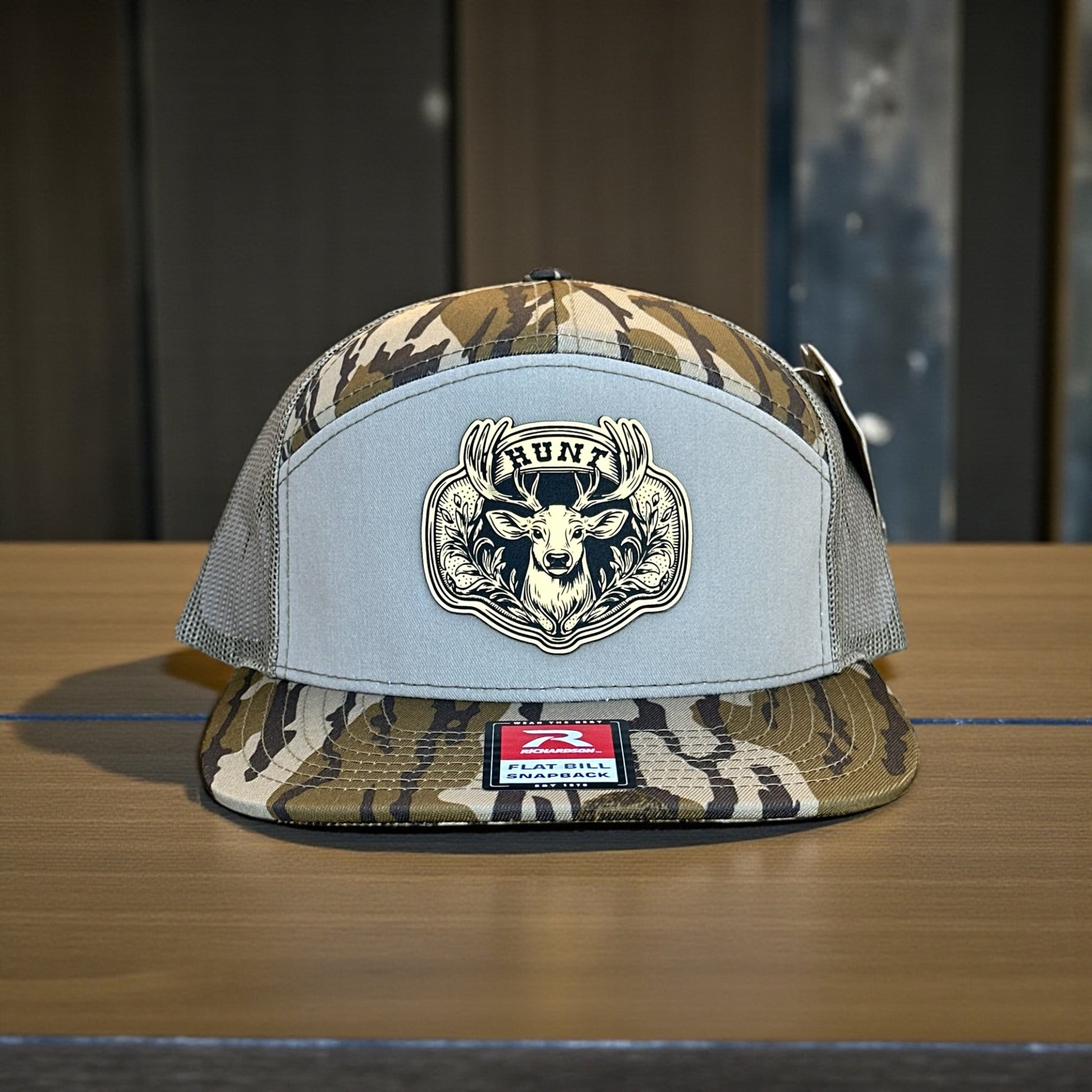 Camouflage trucker cap with a laser engraved deer patch with the text Hunt on a wooden surface with a wooden background