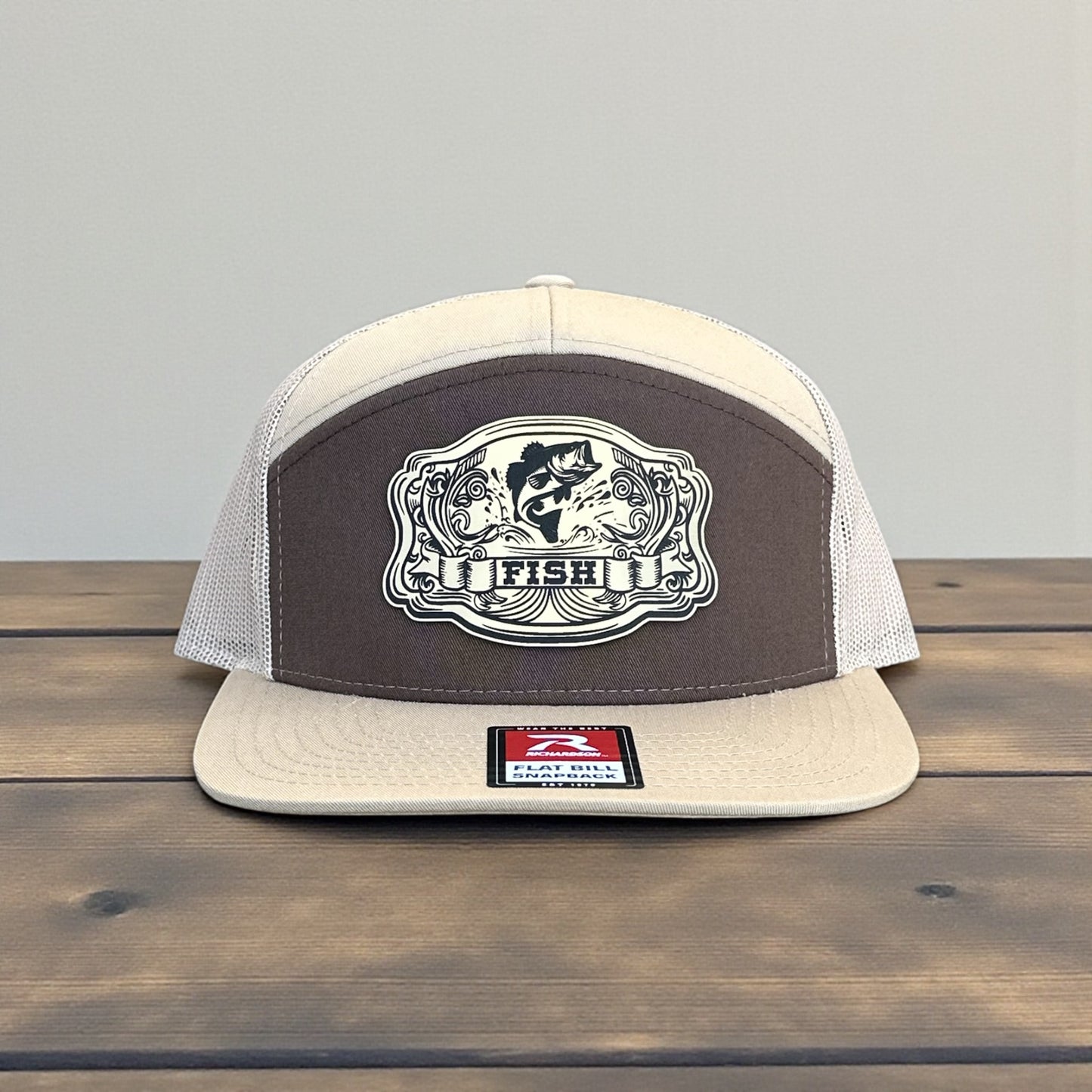 Beige and brown trucker cap with a laser engraved fish patch with the text Fish logo on a wooden surface and grey background