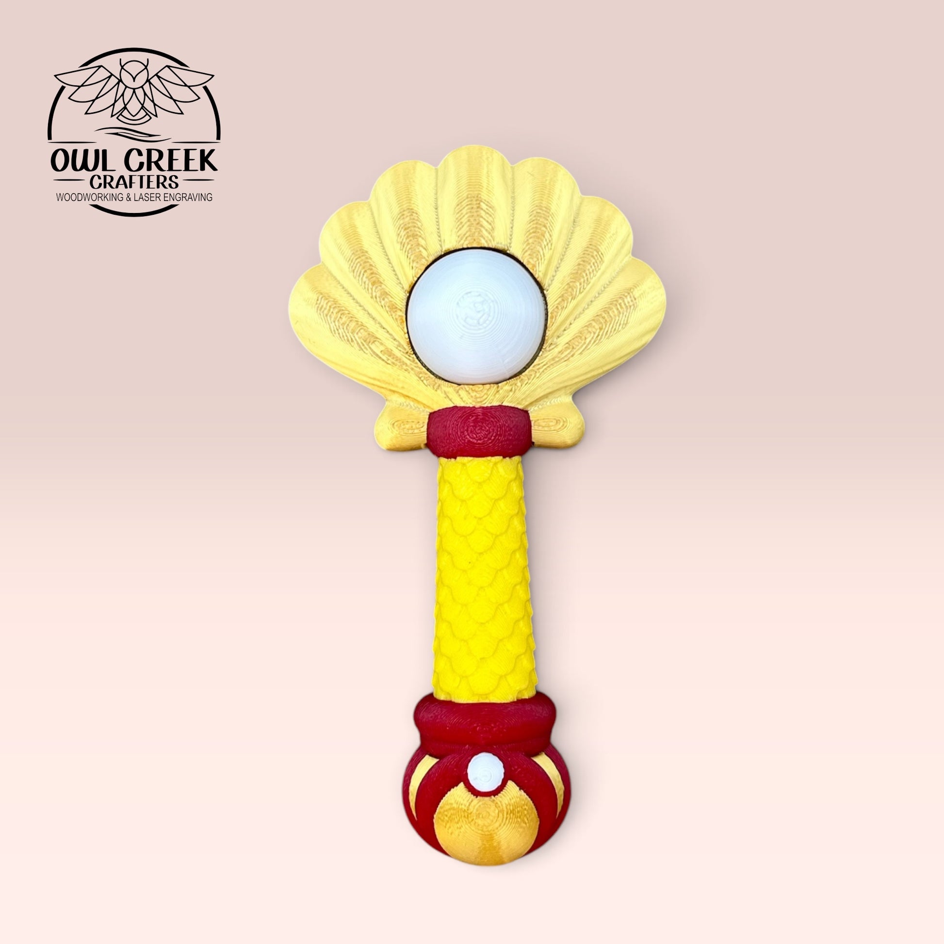 A 3d mermaid clicker wand, featuring a gold shell-shaped top, a yellow textured handle, and red and gold accents, shown on a light pink background with the Owl Creek Crafts logo in the top left corner.