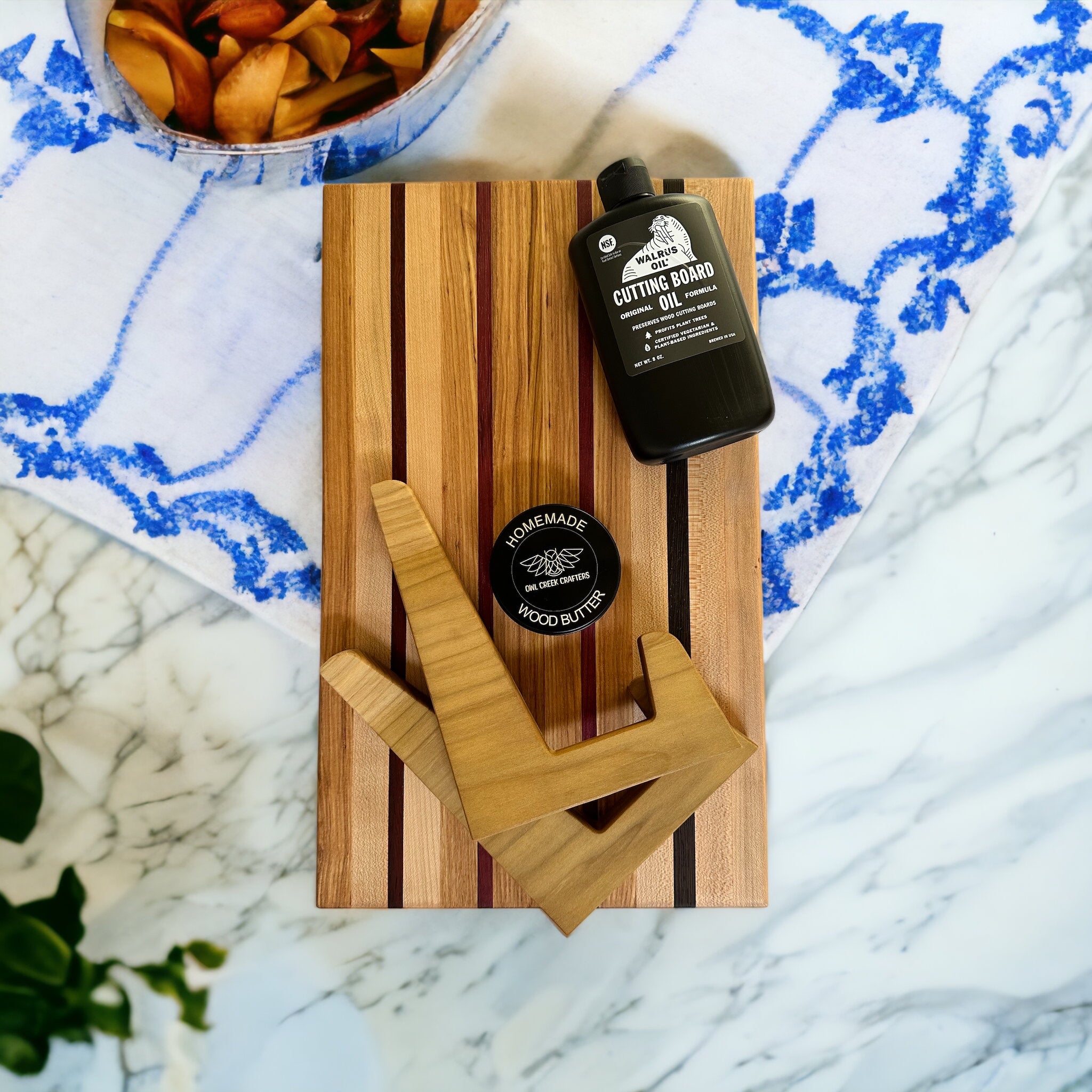 multi-wood cutting board with a bottle of cutting board oil, a can of wood butter, and a pair of wooden cutting board holders, sitting on a blue and white dish towel on a marble countertop with a bowl of fruit and a plant in the background