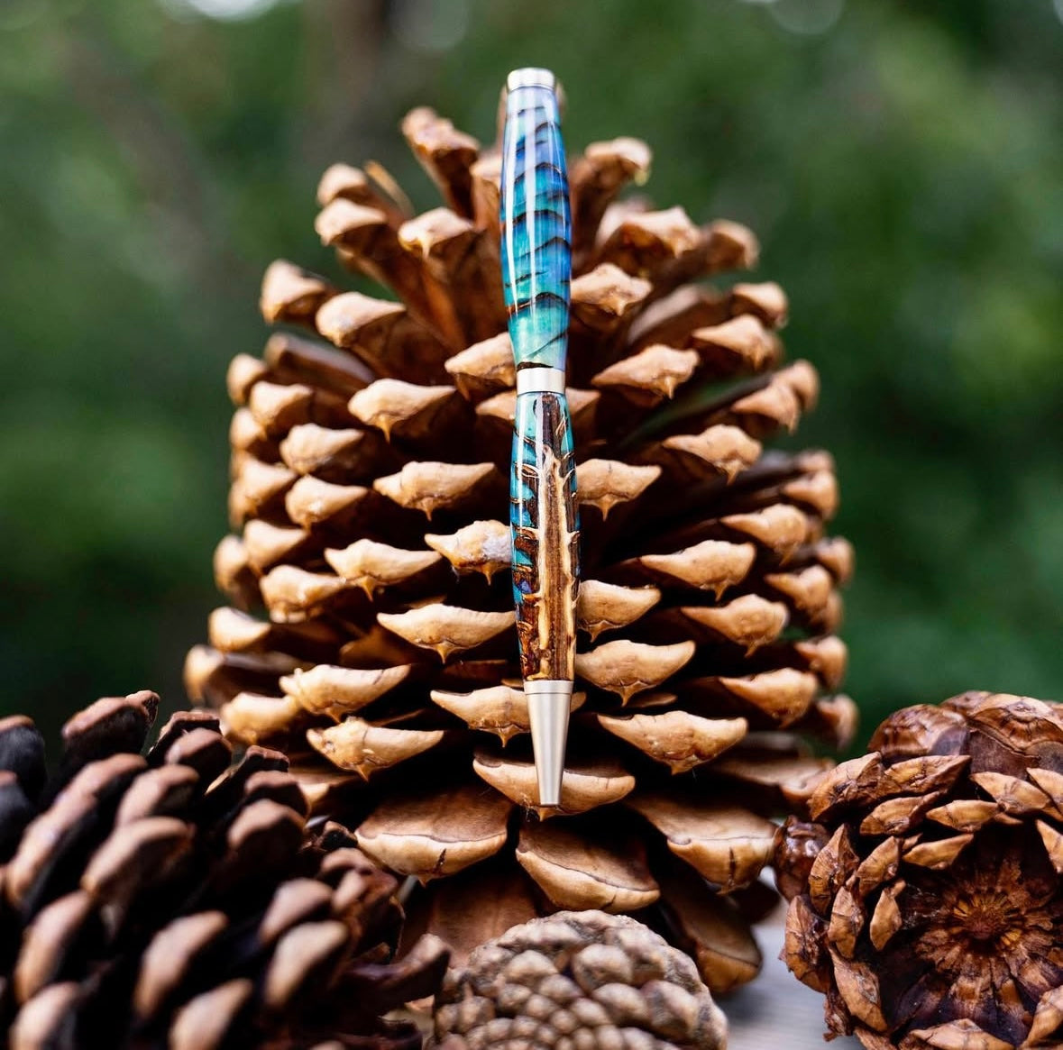 Blue epoxy and pinecone pen with silver accents leaning against pinecones with a green background