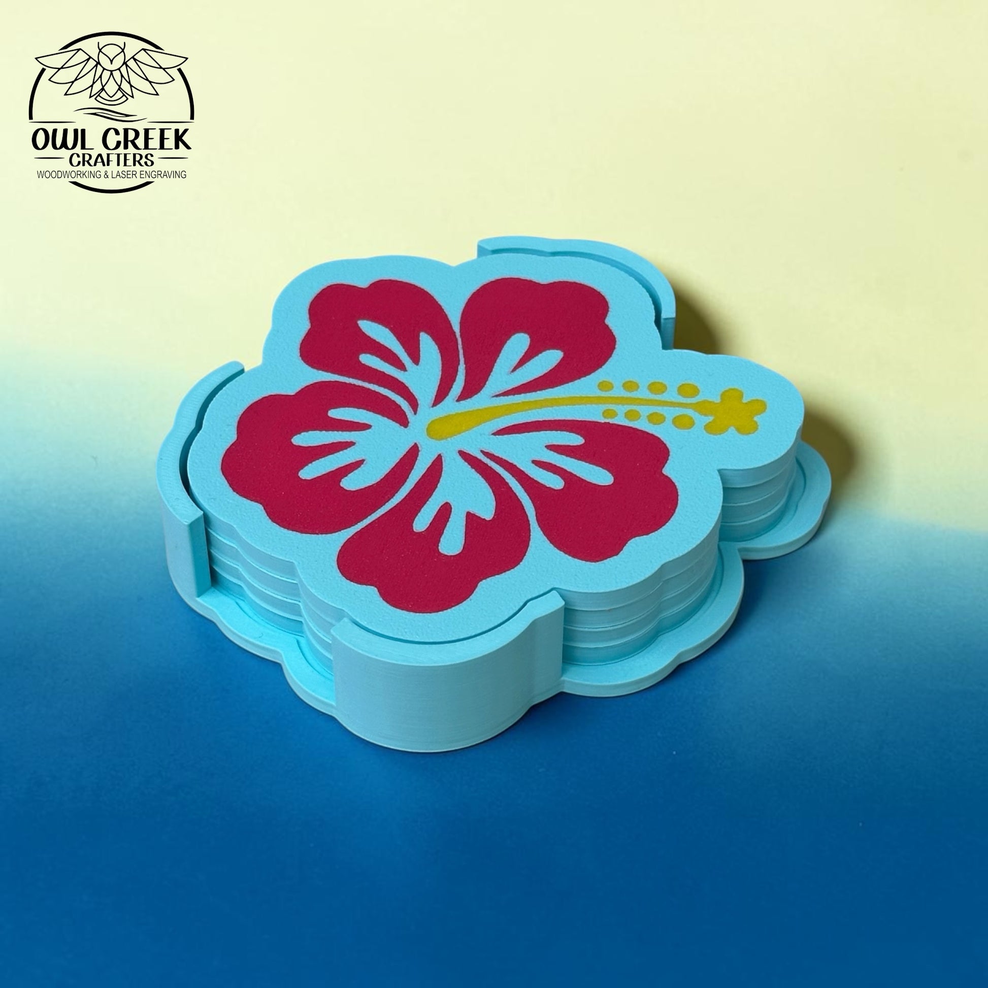 A stack of light blue flower-shaped coasters featuring a large magenta hibiscus flower with a yellow stamen, displayed on a blue surface against a pale yellow background. The Owl Creek Crafters logo is visible in the top-left corner.