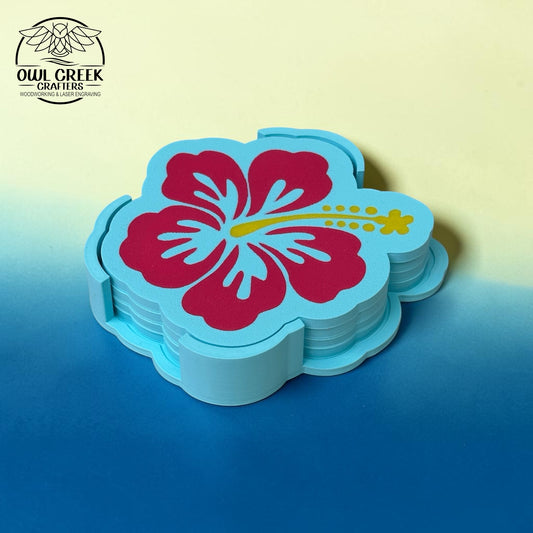 A stack of light blue flower-shaped coasters featuring a large magenta hibiscus flower with a yellow stamen, displayed on a blue surface against a pale yellow background. The Owl Creek Crafters logo is visible in the top-left corner.