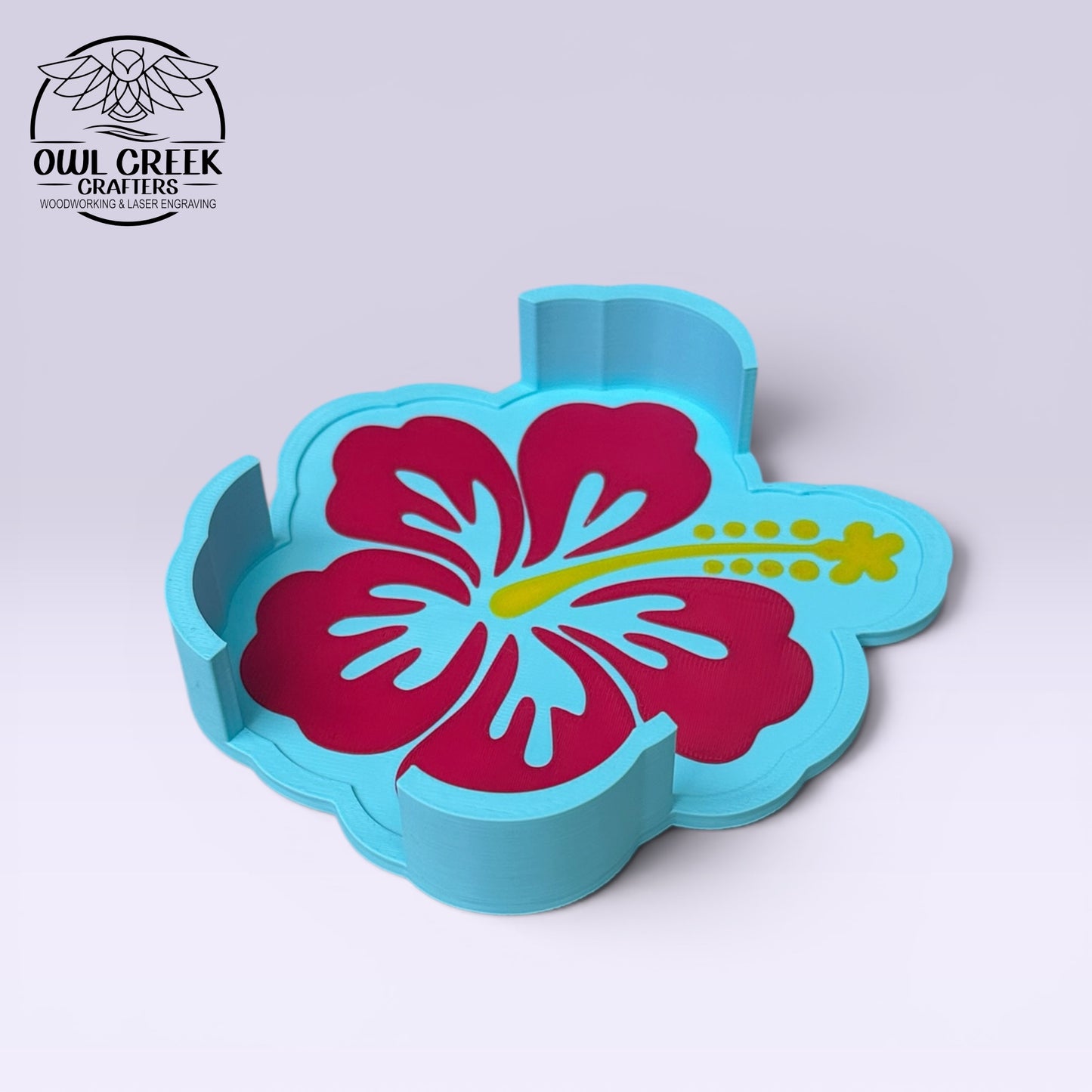 A light blue flower-shaped coaster rack featuring a large magenta hibiscus flower with a yellow stamen, shown on a light gray background. The Owl Creek Crafters logo is visible in the top-left corner.