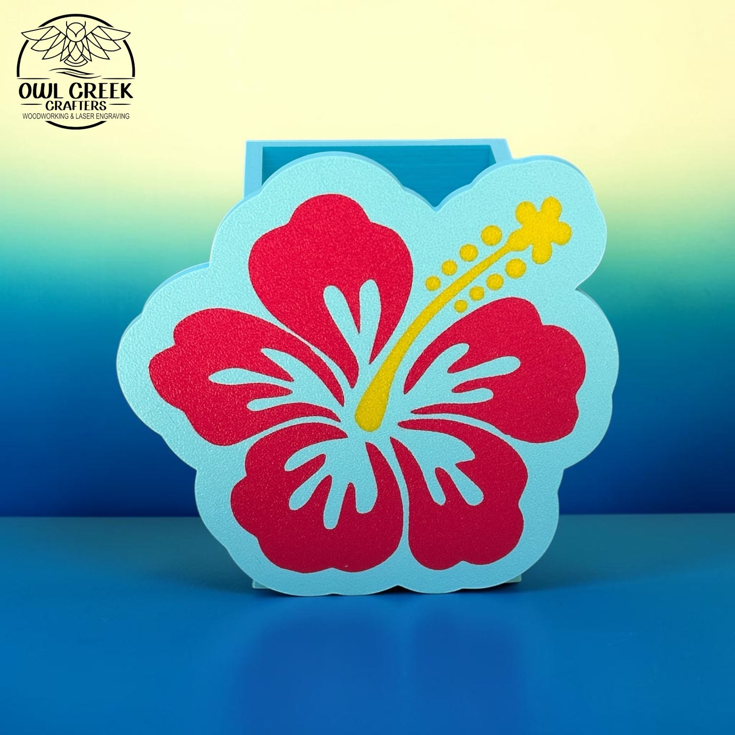 A decorative 3d printed hibiscus flower pencil holder in magenta and light blue with a yellow stamen, displayed on a blue surface against a blue-to-yellow gradient background. The Owl Creek Crafters logo is visible in the top left corner.