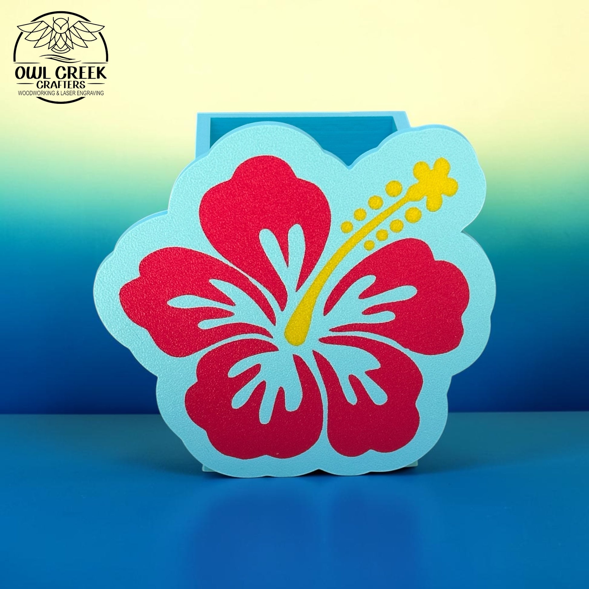 A decorative 3d printed hibiscus flower pencil holder in magenta and light blue with a yellow stamen, displayed on a blue surface against a blue-to-yellow gradient background. The Owl Creek Crafters logo is visible in the top left corner.
