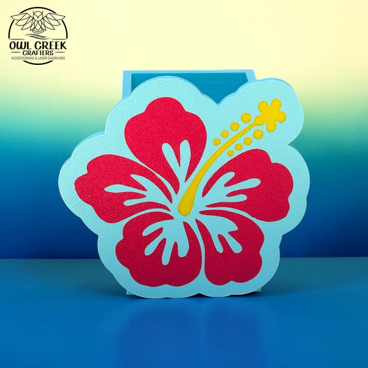 A decorative 3d printed hibiscus flower pencil holder in magenta and light blue with a yellow stamen, displayed on a blue surface against a blue-to-yellow gradient background. The Owl Creek Crafters logo is visible in the top left corner.