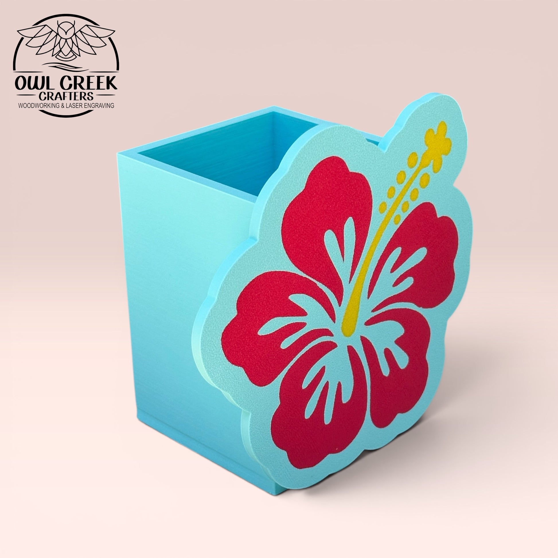 A decorative 3d printed hibiscus flower pencil holder in magenta and light blue with a yellow stamen, displayed on a light pink background. The Owl Creek Crafters logo is visible in the top left corner.
