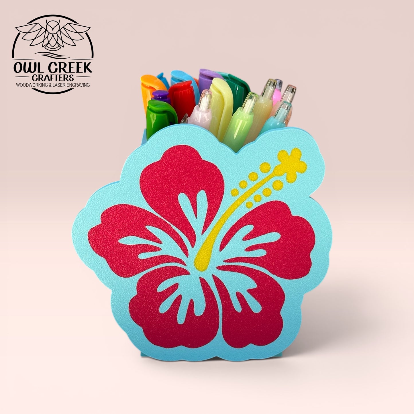A decorative 3d printed hibiscus flower pencil holder in magenta and light blue with a yellow stamen, holding several colorful pens displayed on a light pink background. The Owl Creek Crafters logo is visible in the top left corner.
