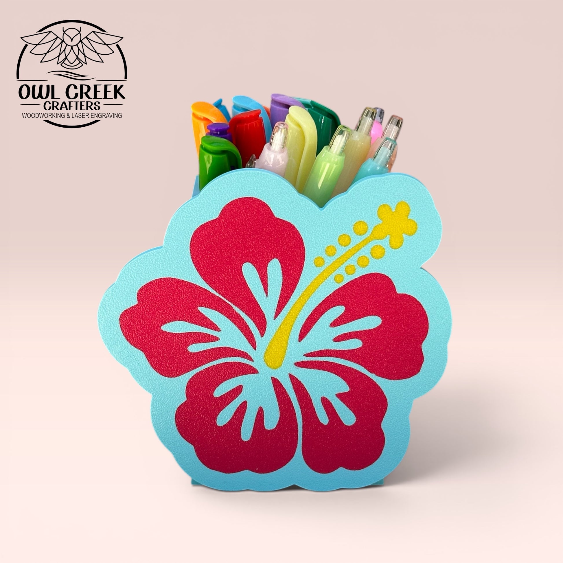 A decorative 3d printed hibiscus flower pencil holder in magenta and light blue with a yellow stamen, holding several colorful pens displayed on a light pink background. The Owl Creek Crafters logo is visible in the top left corner.