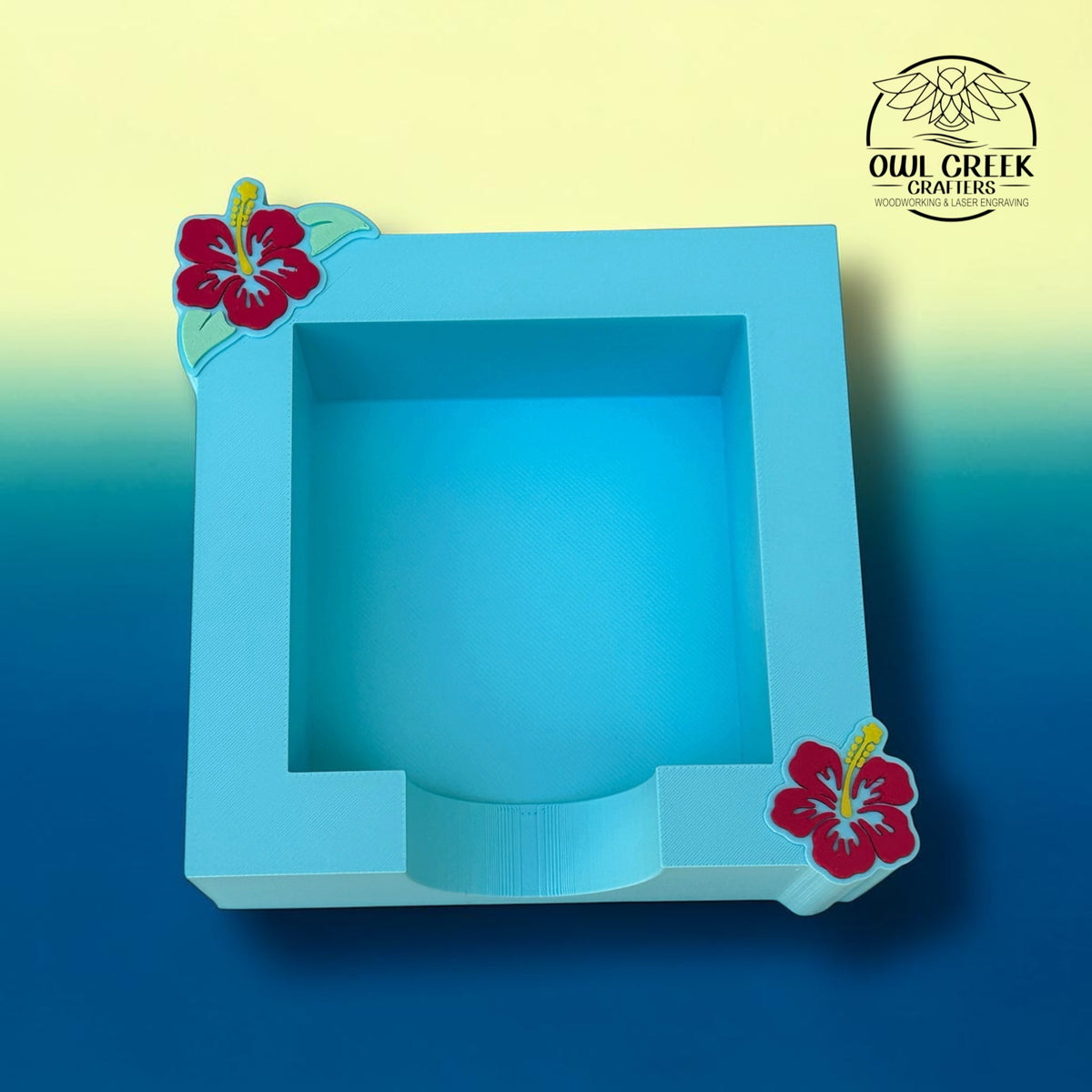 A 3d printed light blue sticky note holder with two magenta hibiscus flowers in the top left and bottom right corners. On a blue to yellow gradient background with the Owl Creek Crafters logo in the top right corner.