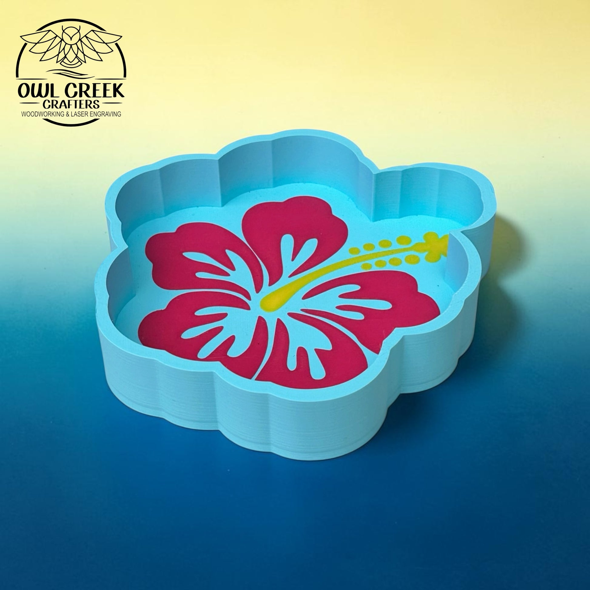 A light blue flower-shaped trinket tray with a pink hibiscus flower and yellow stamen design printed inside, shown on a blue-to-yellow gradient background. The Owl Creek Crafters logo is visible in the top-left corner.
