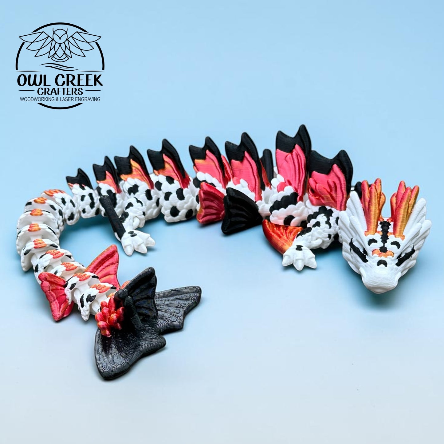 A 3d printed dragon figurine by Owl Creek Crafters, featuring a white body with black spots, red and orange wing accents, and a black tail, displayed on a light blue background.