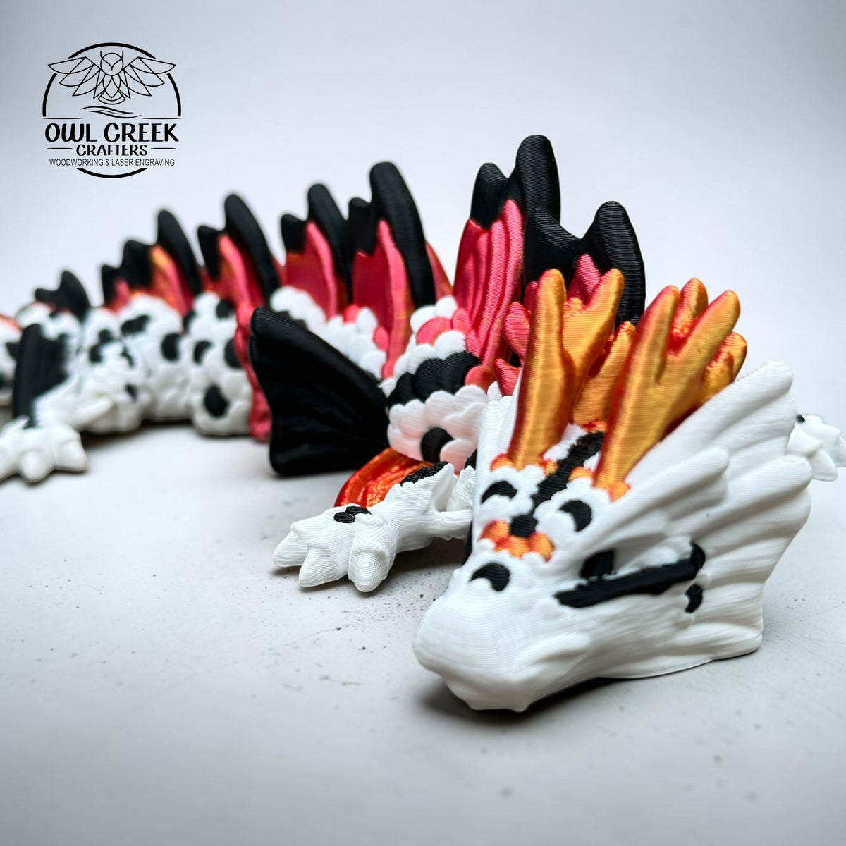A close up of a 3d printed dragon figurine by Owl Creek Crafters, featuring a white body with black spots, red and orange wing accents, and a black tail, displayed on a light blue background.