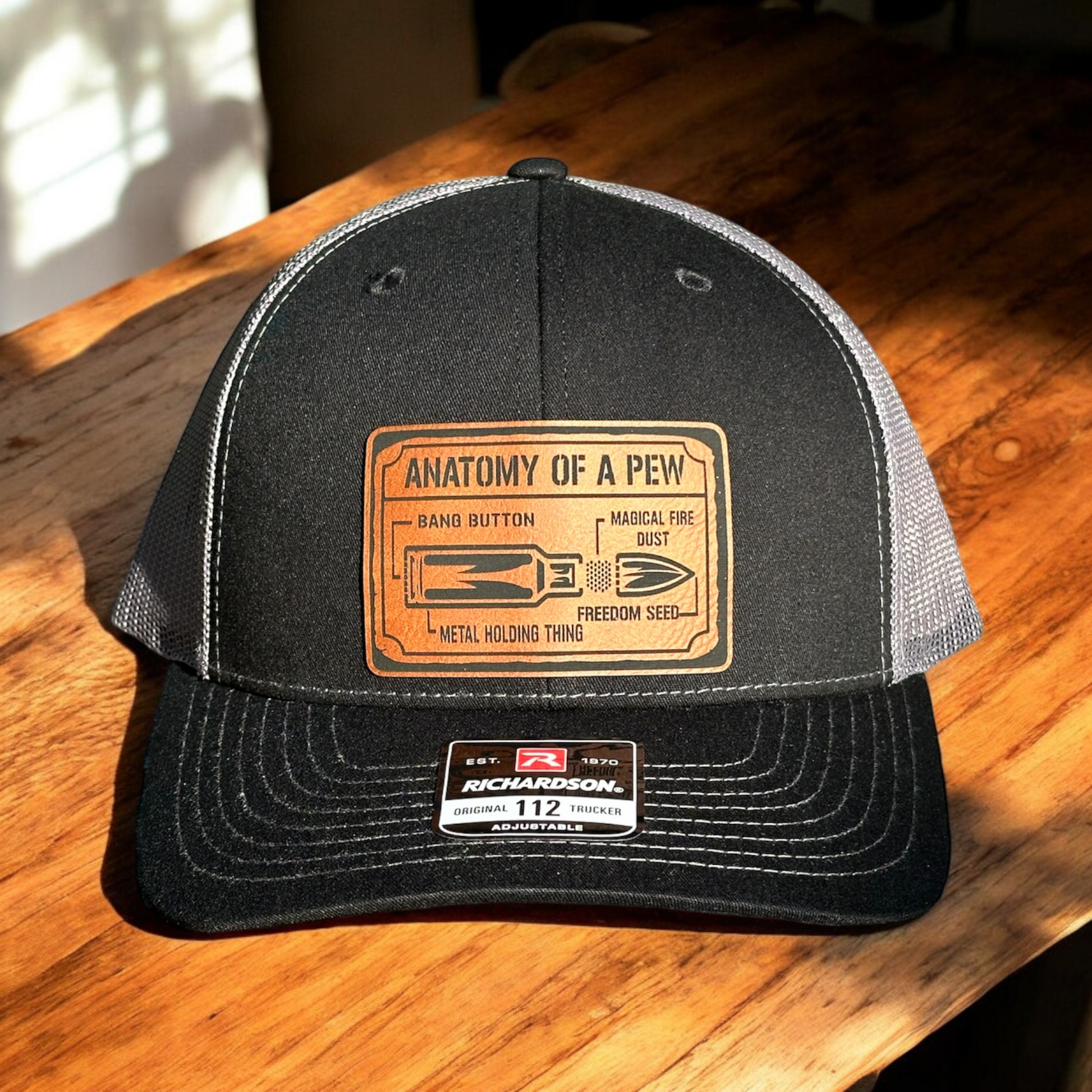 A black trucker hat with a gray mesh back and a leatherette patch on the front featuring text 'anatomy of a pew' and a graphic, sitting on a wooden surface.