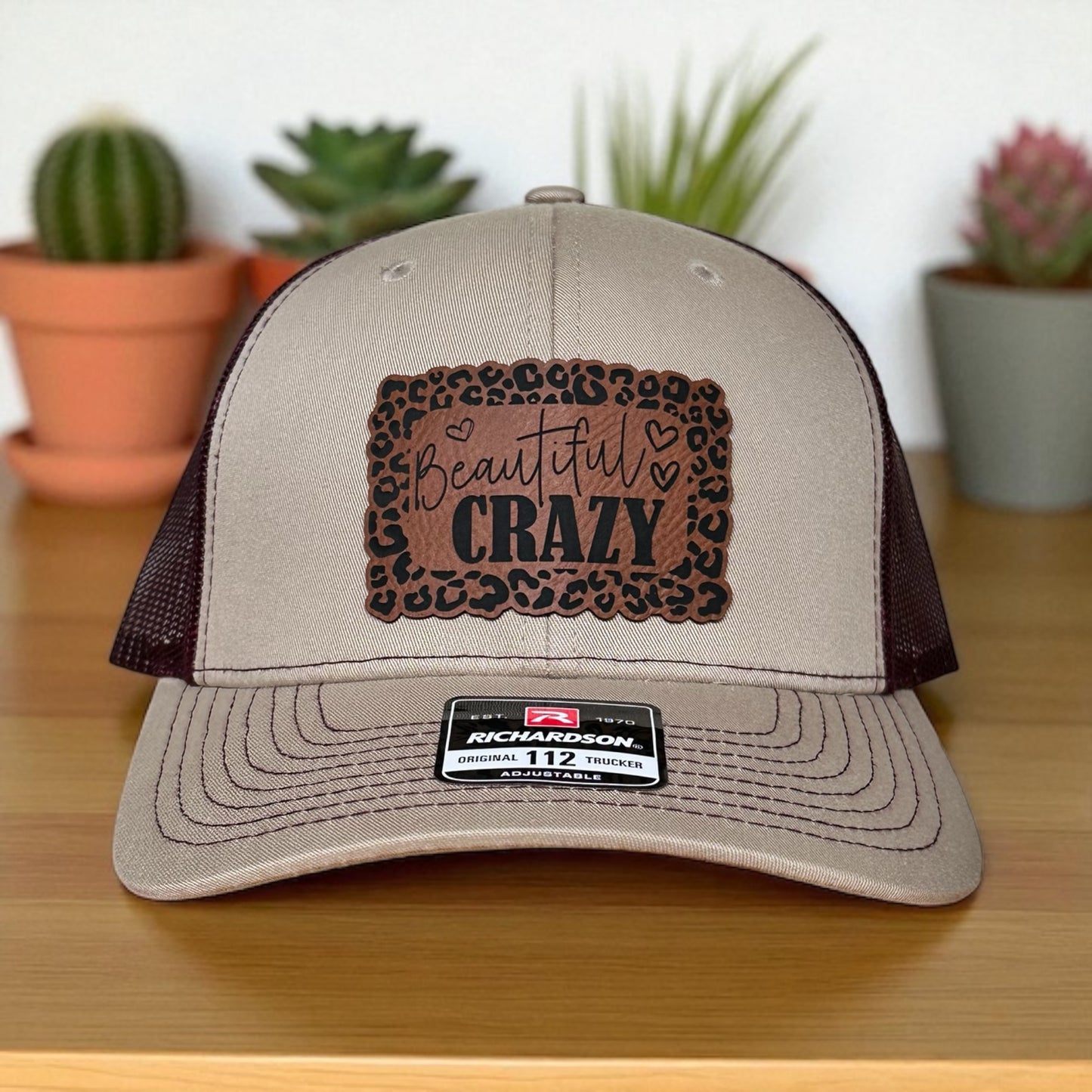 Beige trucker cap with burgandy mesh back, and a leopard print leatherette patch with the text 'Beautiful Crazy', on a wooden surface with plants in the background