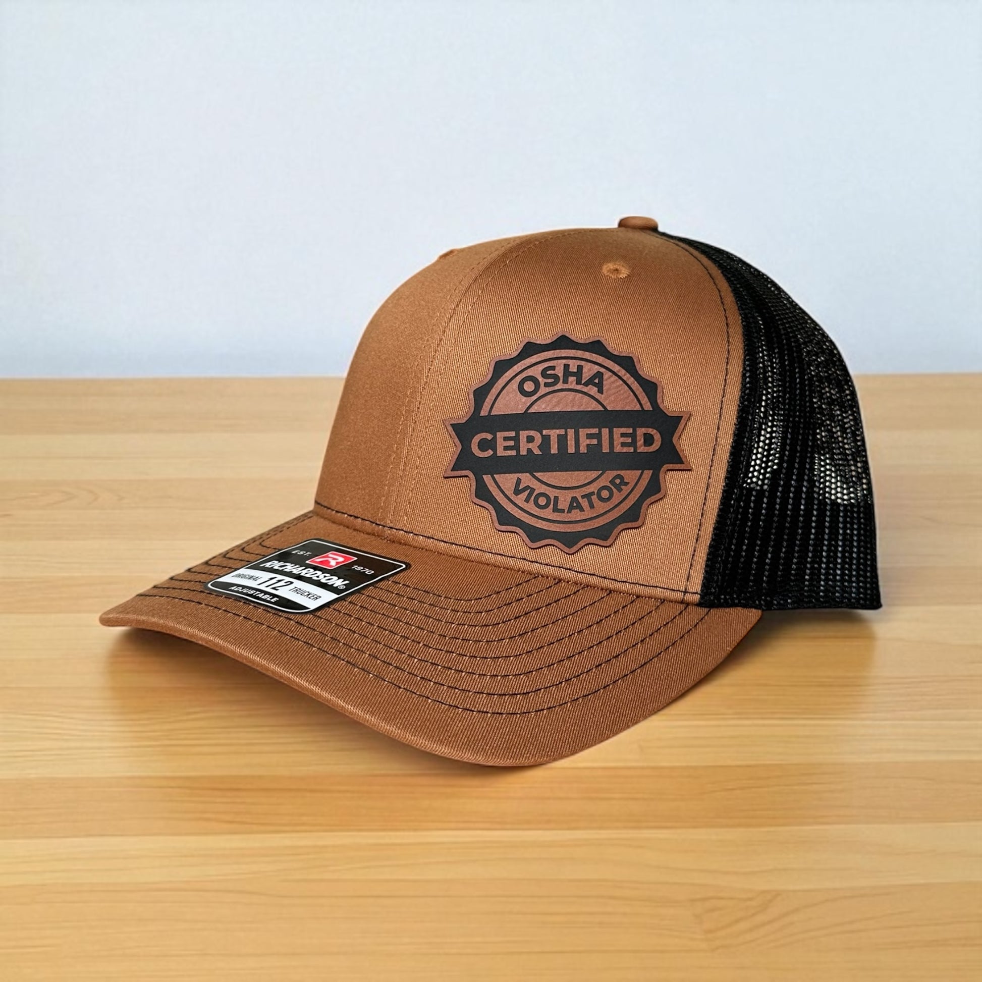 Brown trucker cap with black mesh back and a leatherette patch with the text 'OSHA Certified Violator', on a wooden surface with a grey background