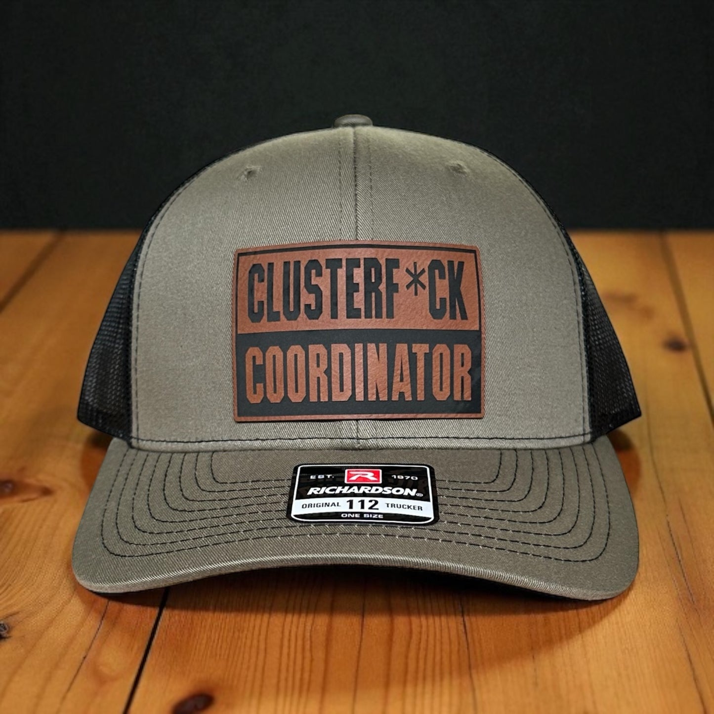 Green trucker cap with black mesh back and a leatherette patch with the text 'Clusterf*ck Coordinator', on a wooden surface with a black background