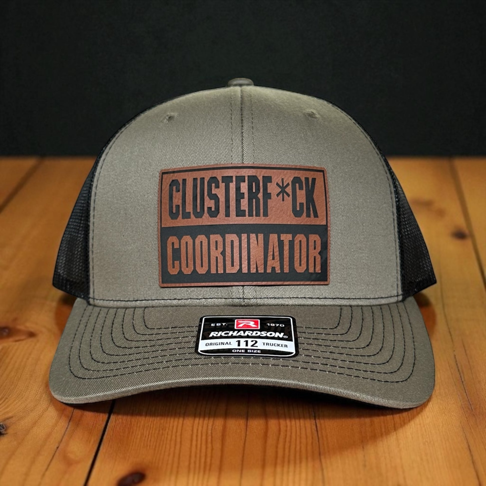 Green trucker cap with black mesh back and a leatherette patch with the text 'Clusterf*ck Coordinator', on a wooden surface with a black background