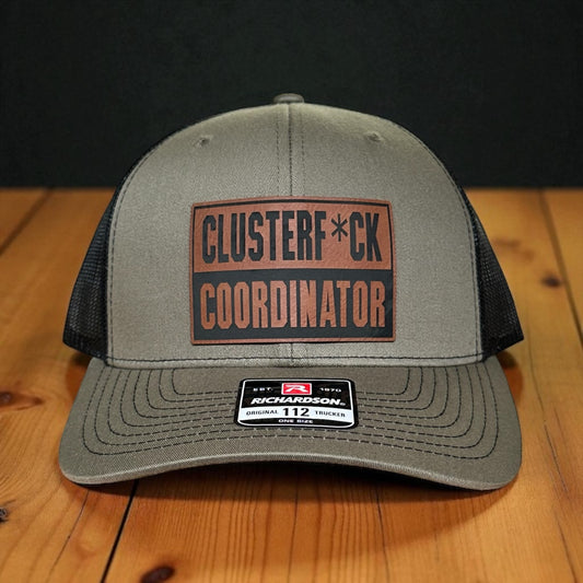 Green trucker cap with black mesh back and a leatherette patch with the text 'Clusterf*ck Coordinator', on a wooden surface with a black background