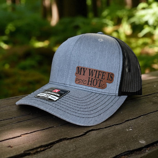 Gray trucker cap with a leatherette patch on the side with the text 'My Wife is psycHOTic' on a wooden surface with a blurred natural woods background