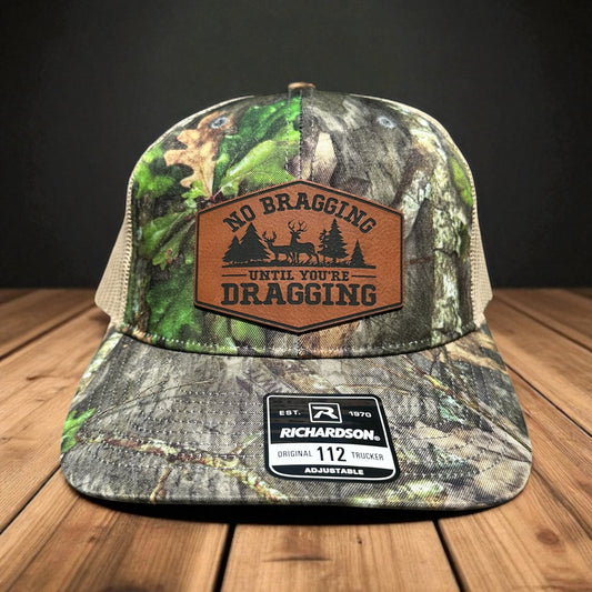 Camouflage trucker hat with leatherette patch and the text 'No Bragging until you're dragging' and a deer scene, on a wooden surface and a dark gray background