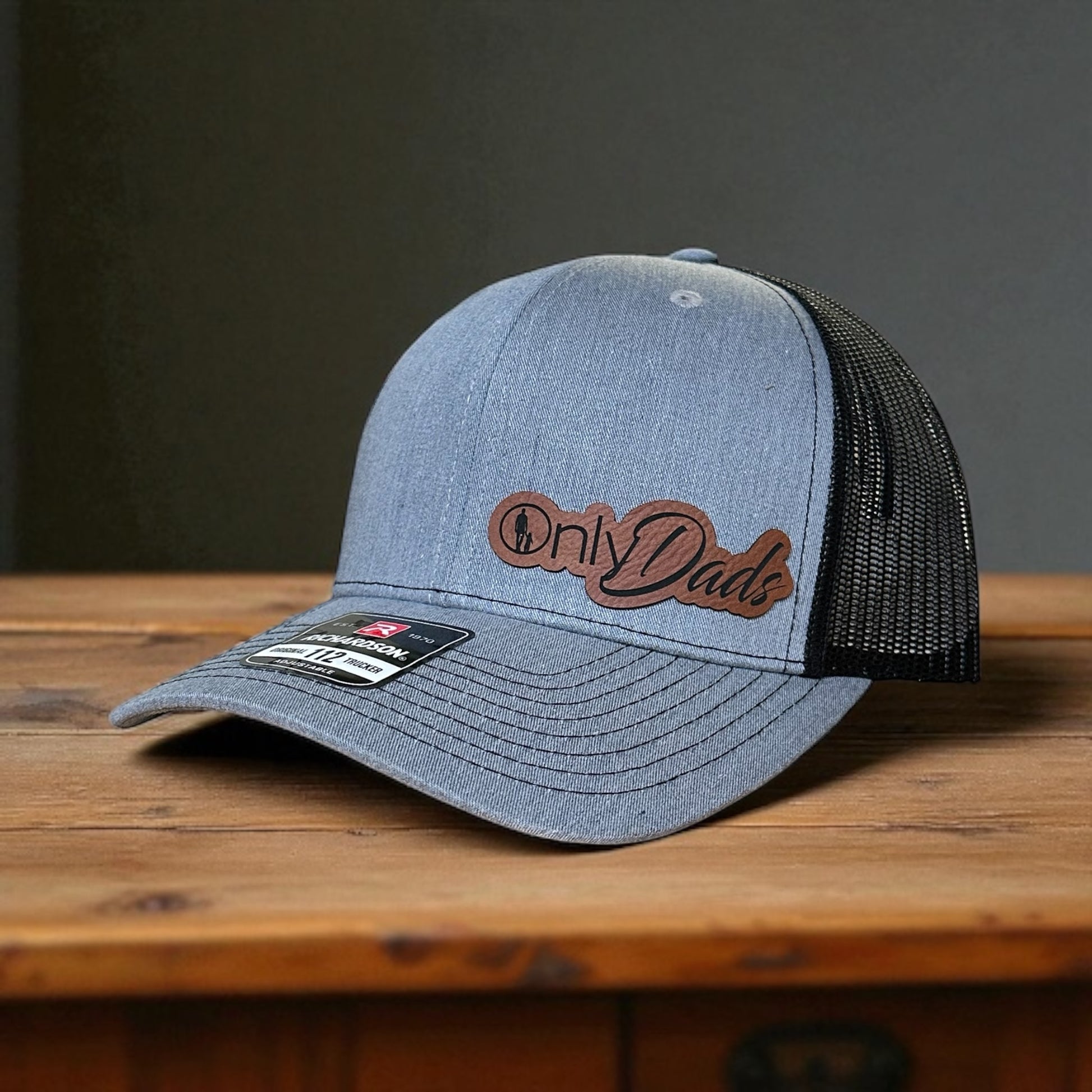 Gray trucker hat with black mesh back, and a leatherette patch with the text  'Only Dads', on a wooden surface with a gray background