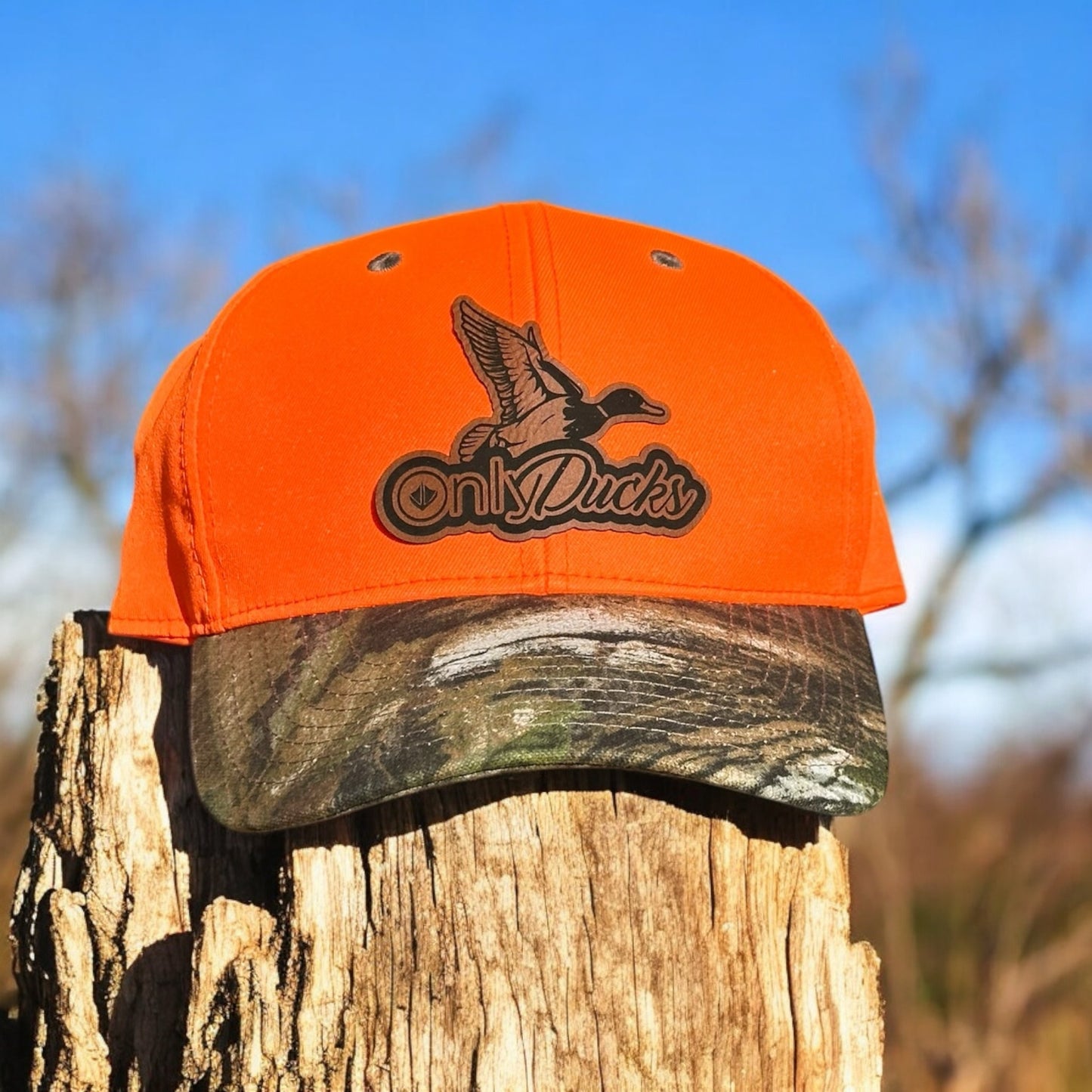 Two-tone cap with an orange top and a camoflage bill, with a leatherette patch of a duck design with the text 'Only Ducks', on a wooden surface with a natural outdoors in the background