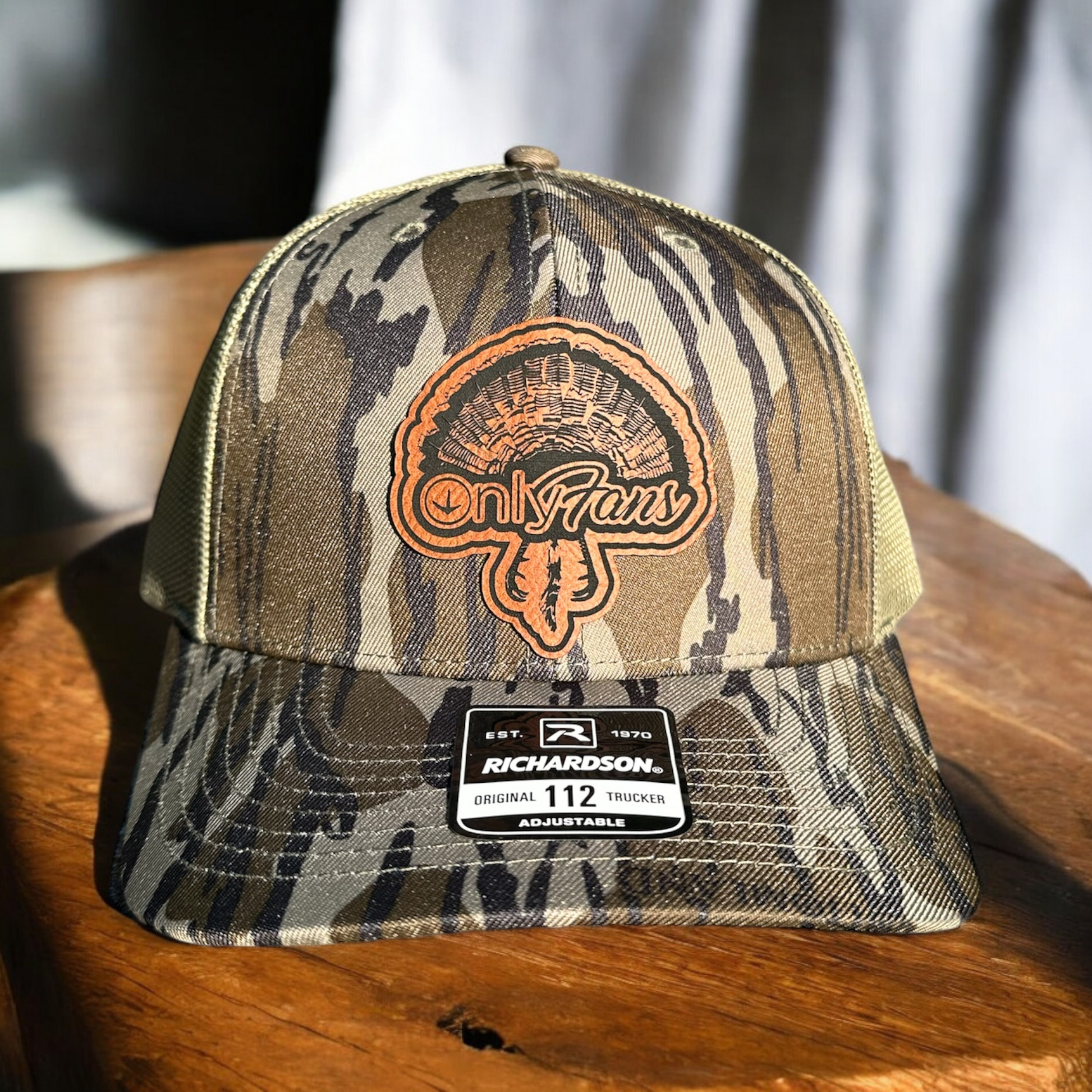 Camoflage trucker cap with a leatherette patch with the text 'Only Fans' and a turkey fan design, on a wooden surface with a linen background