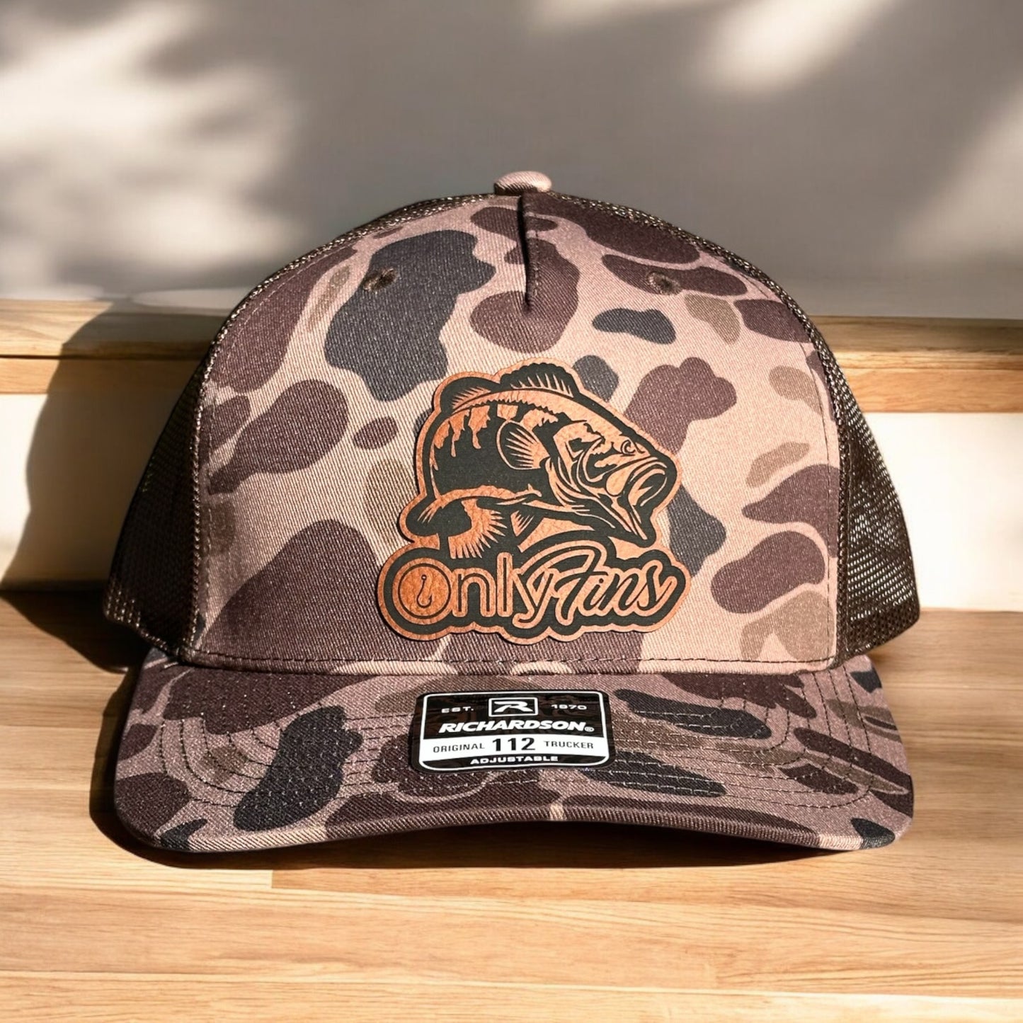 Brown camoflage trucker hat with a laser engraved leatherette patch with a fish design and a text 'Only Fins', on a wooden surface with a beige background