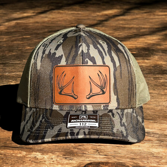 Green camoflage trucker hat with a laser engraved leatherette patch and a deer antler rack design, on a wooden surface