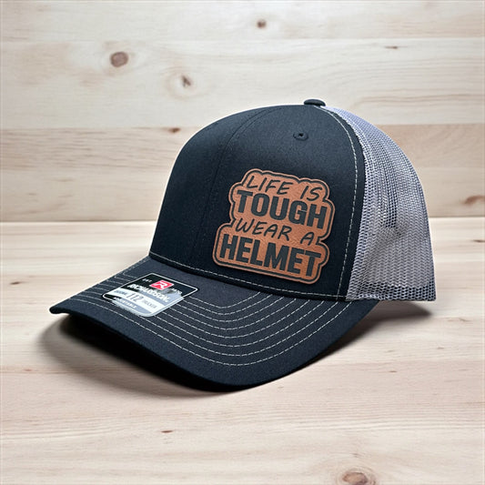 Black trucker cap with grey mesh back and a leatherette patch on the side with the test 'Life is Tough, Wear a Helmet', on a wooden surface and wooden background
