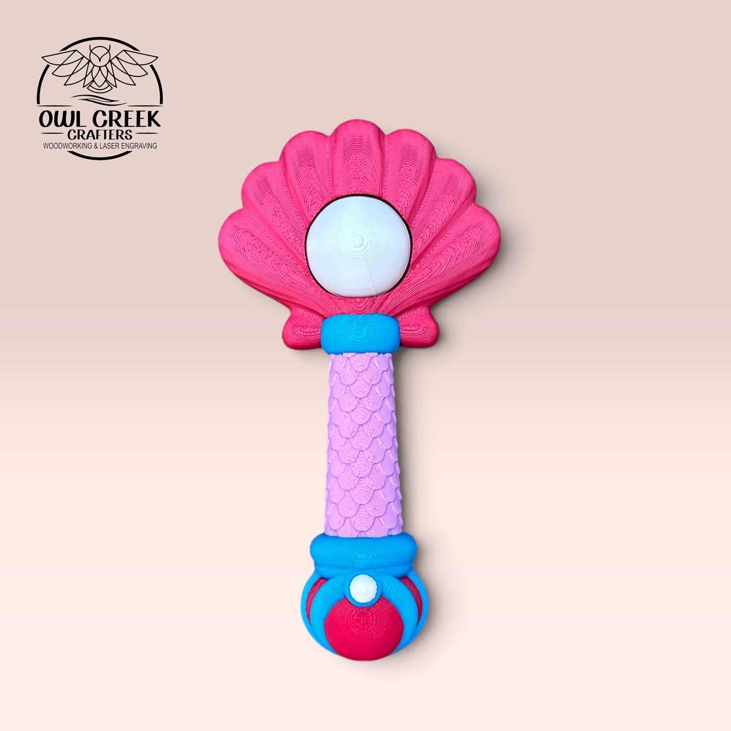 A 3d mermaid clicker wand, featuring a pink shell-shaped top, a purple textured handle, and blue and pink accents, shown on a light pink background with the Owl Creek Crafts logo in the top left corner.