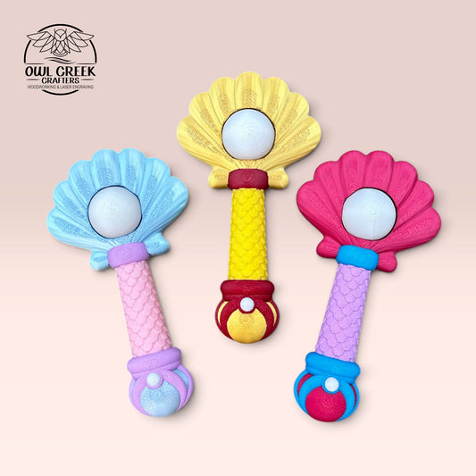 Three colorful shell-shaped 3d printed mermaid clicker toys arranged vertically on a light peach background. The left toy is light blue and pink, the center toy is gold and red, and the right toy is pink and blue. All mermaid clicker toys have a white pearl in the middle that clicks. There is a Owl Creek Crafters logo in the top left corner.