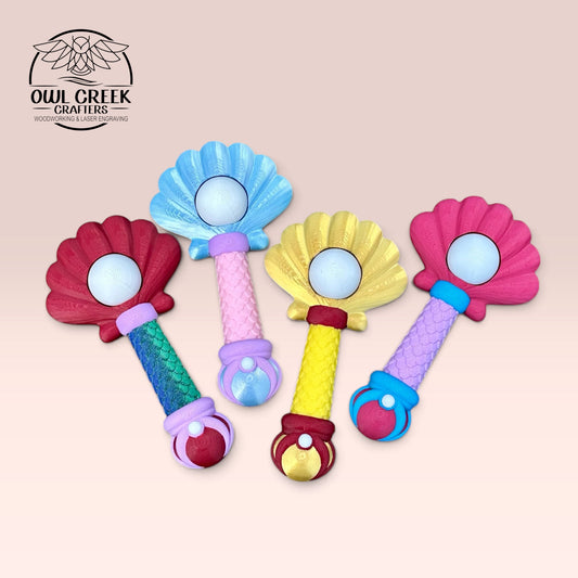 Four colorful sea shell wand toys arranged on a light beige background. The toys have shell-shaped tops in red, blue, yellow, and pink, with textured handles in green, pink, yellow, and purple, and round white "pearl" at the center that clicks. On a beige background with the 'Owl Creek Crafters' Logo in the top left corner.