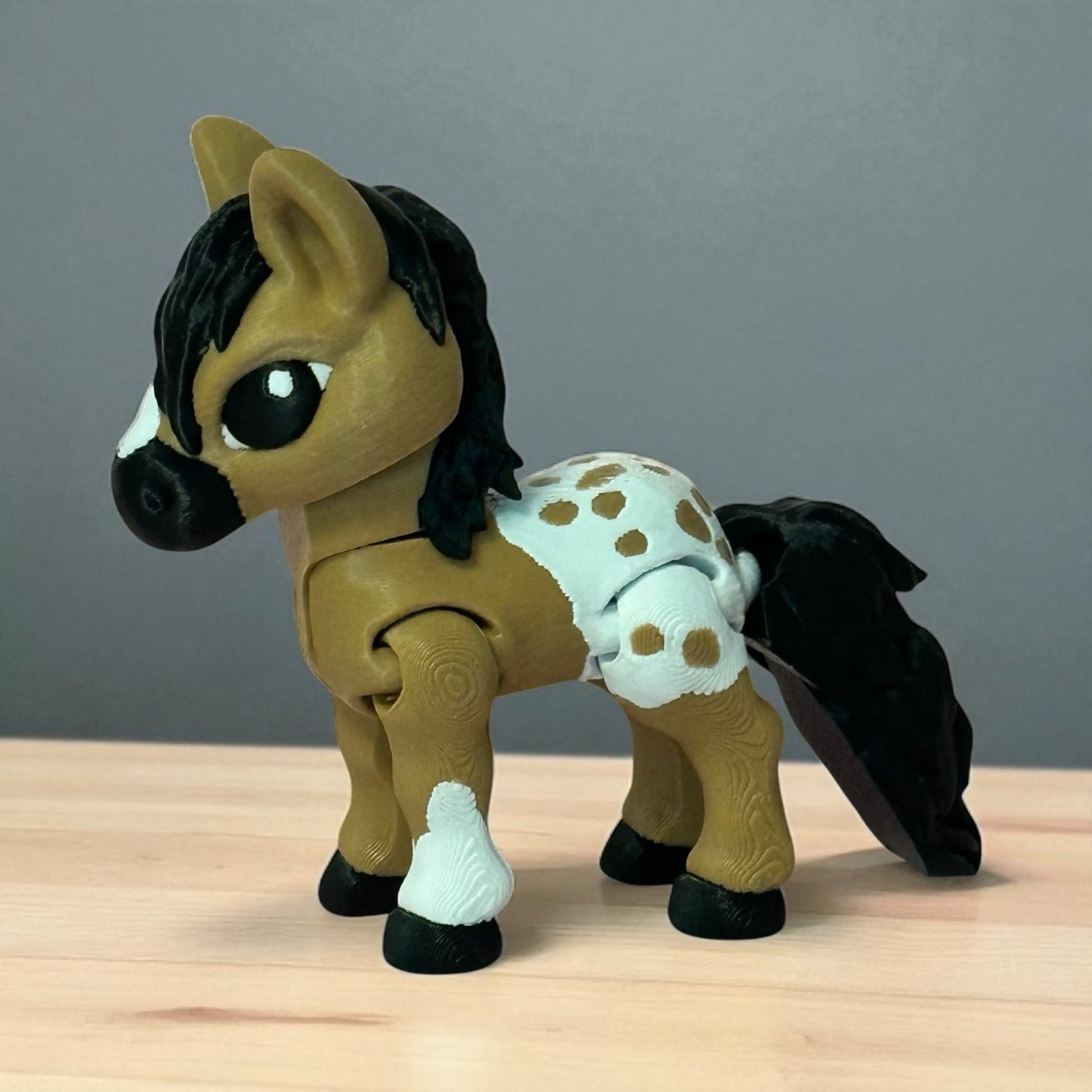 3D printed toy horse with a spotted blanket and white leg on a wooden surface and gray background