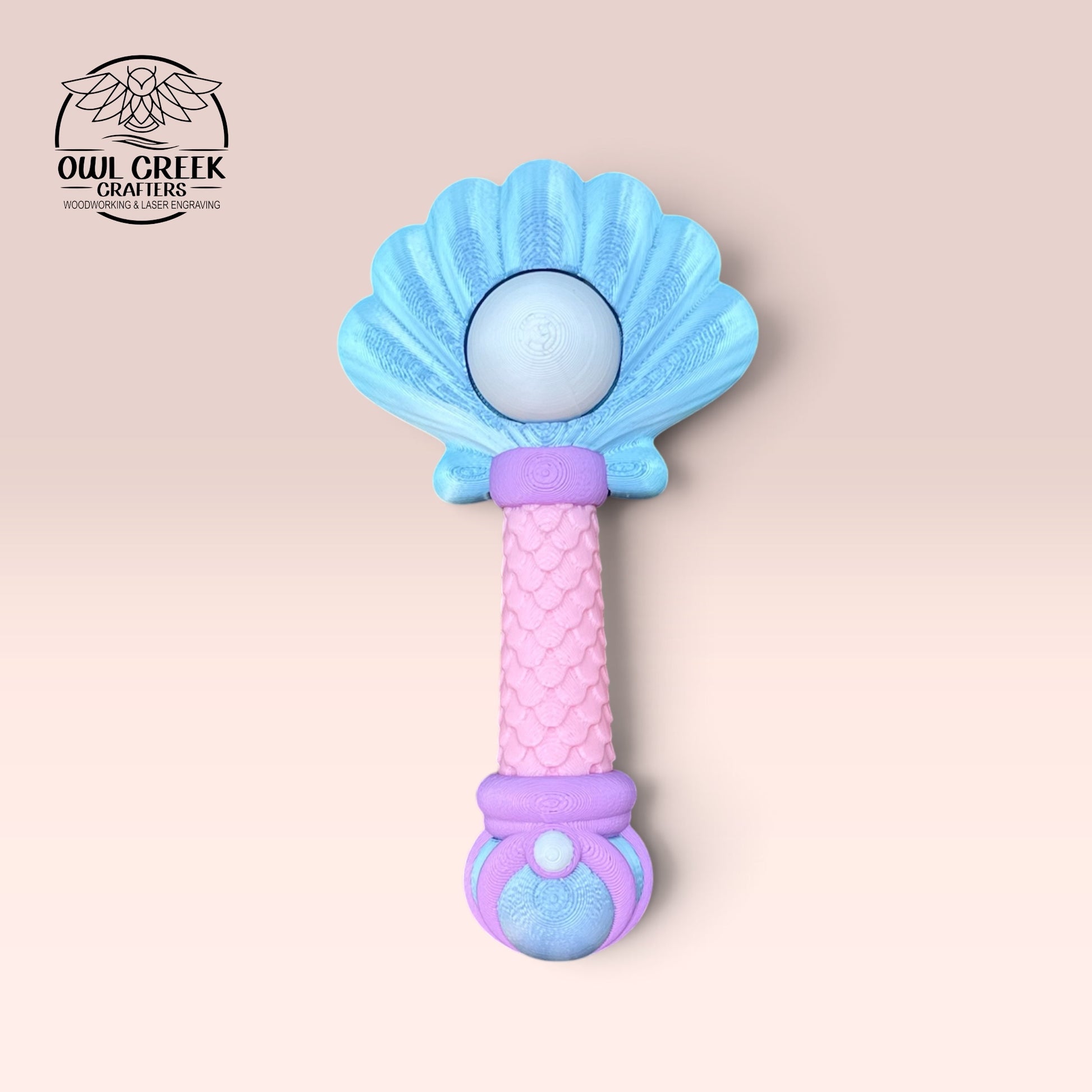 A 3d mermaid clicker wand, featuring a blue shell-shaped top, a pink textured handle, and purple and pink accents, shown on a light pink background with the Owl Creek Crafts logo in the top left corner.