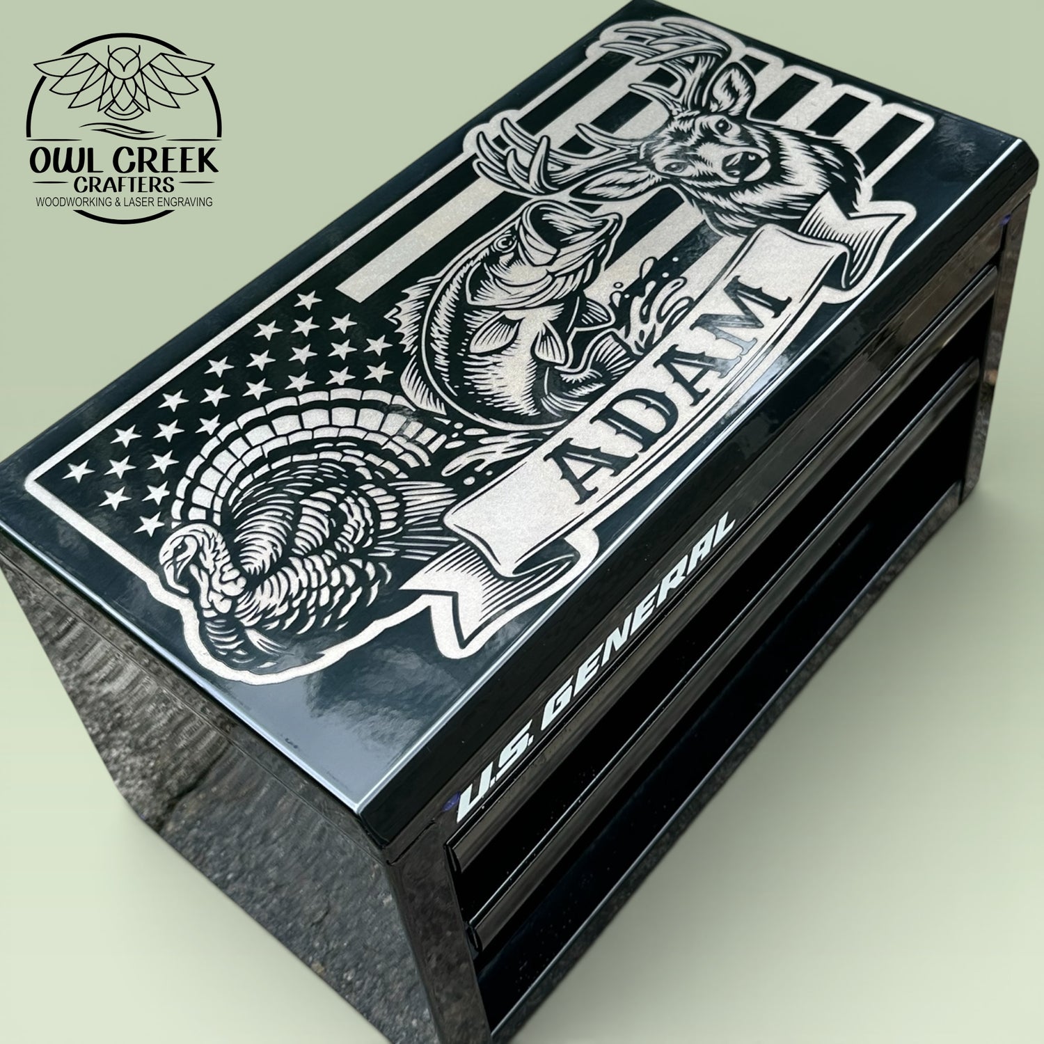 A black metal mini toolbox with a custom engraving on the top with an image of an American flag, a turkey, a bass fish, and a deer with a personalized name, on a green background withe the Owl Creek Crafters Logo in the corner