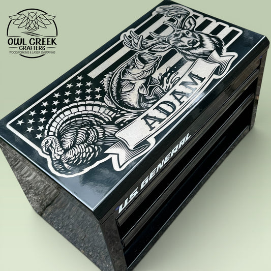 A black metal mini toolbox with a custom engraving on the top with an image of an American flag, a turkey, a bass fish, and a deer with a personalized name, on a green background withe the Owl Creek Crafters Logo in the corner