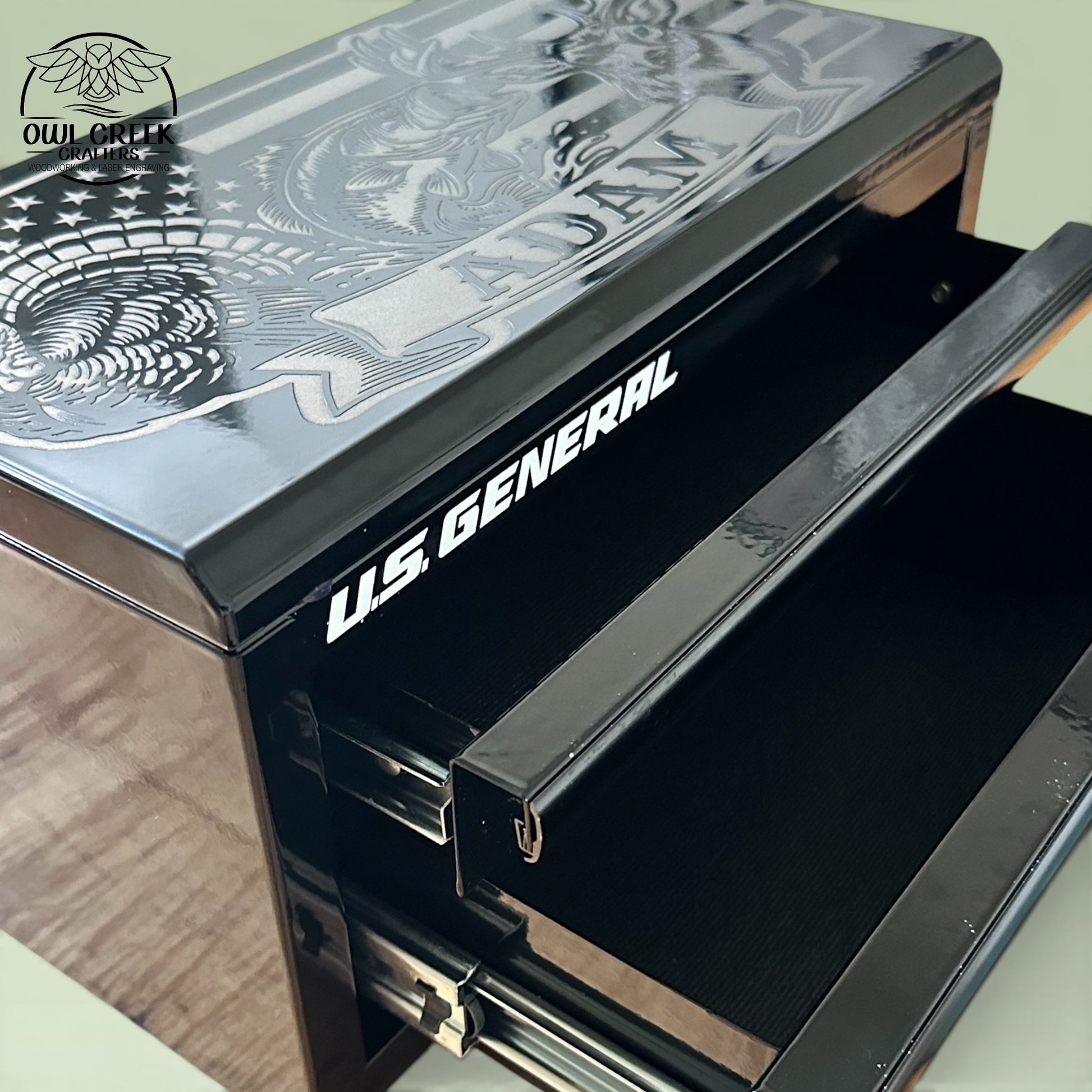 A black metal mini toolbox with a custom engraving on the top with an image of an American flag, a turkey, a bass fish, and a deer with a personalized name, and two drawers open on the toolbox, on a green background withe the Owl Creek Crafters Logo in the corner