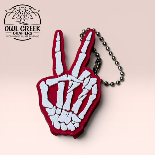 Magenta and white skeleton hand keychain making a peace sign on a beige background with Owl Creek Crafters Logo